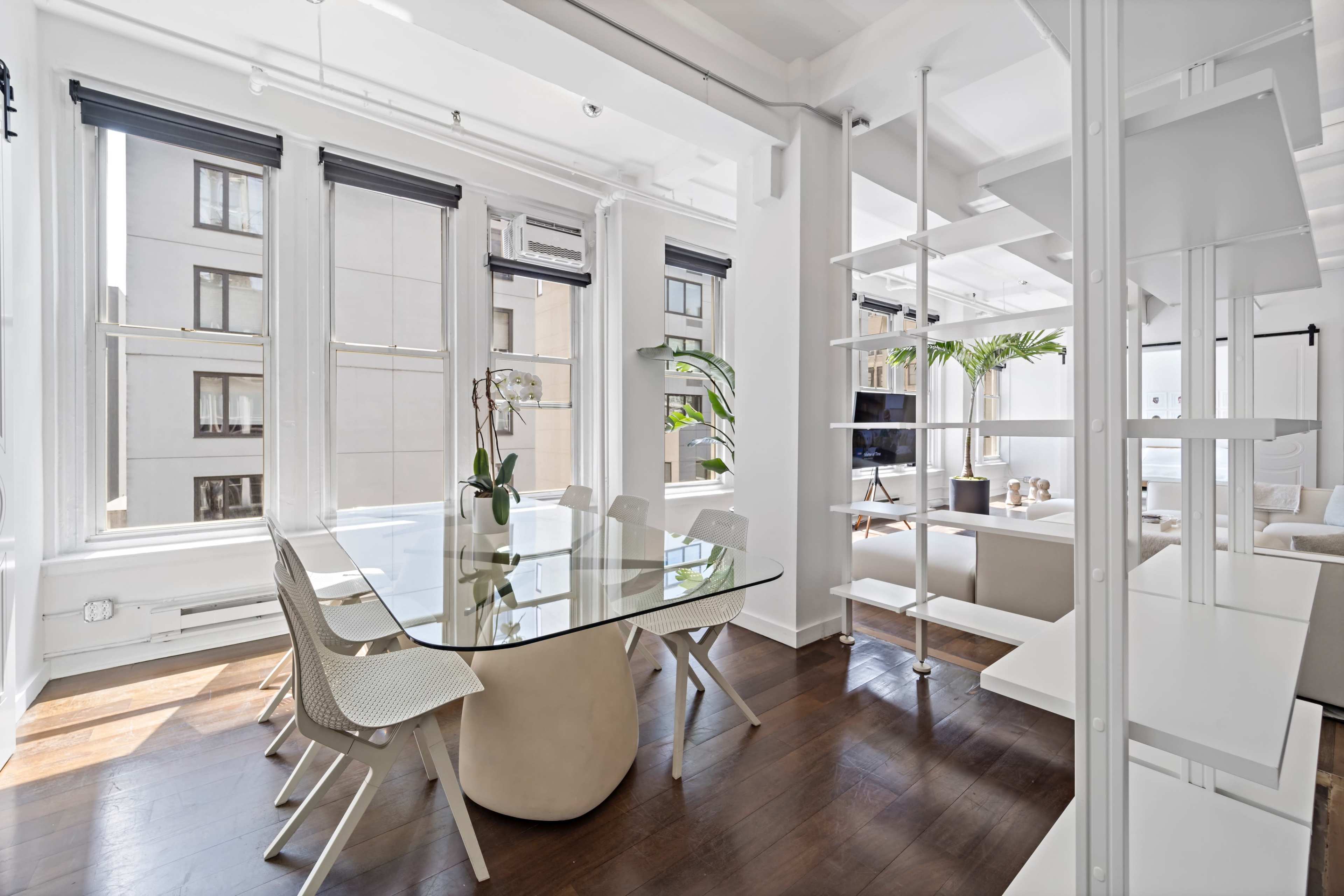 A bright, modern dining area with a glass table surrounded by white chairs and large windows letting in natural light.