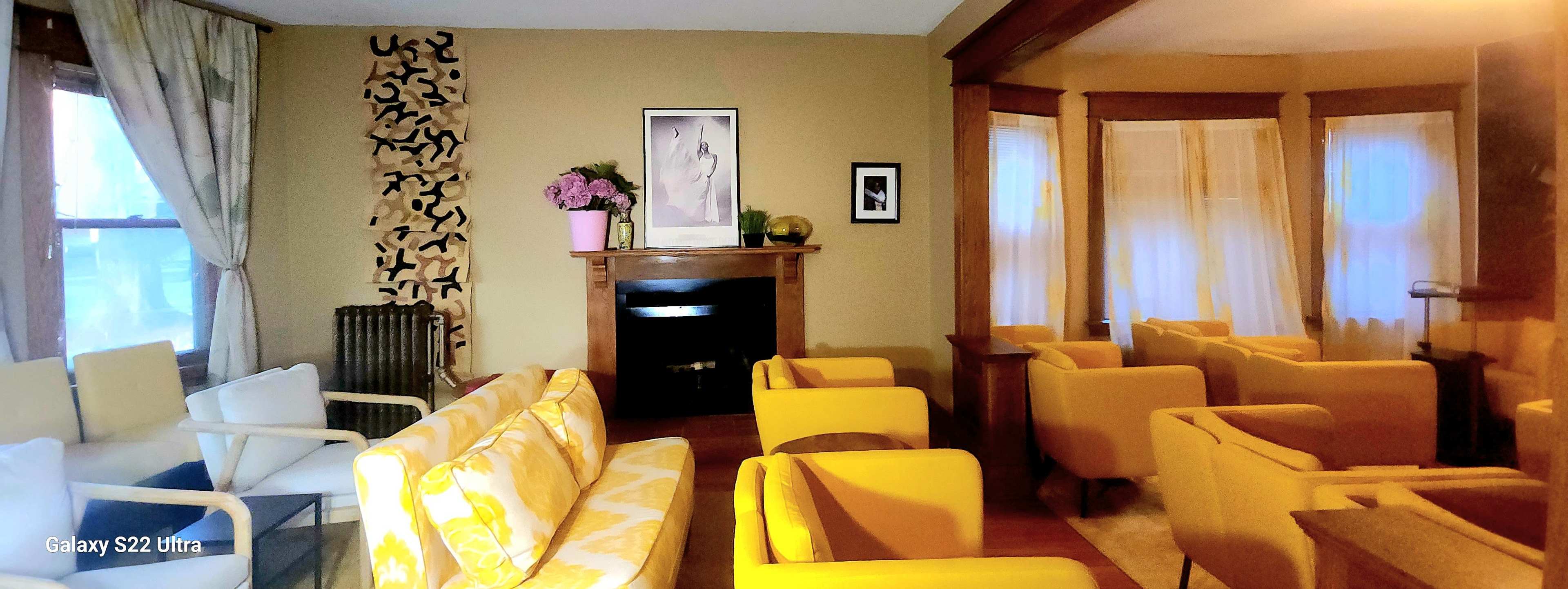 The image shows a living room with a fireplace, yellow upholstered chairs, a white sofa with yellow patterns, and large windows allowing natural light to enter.