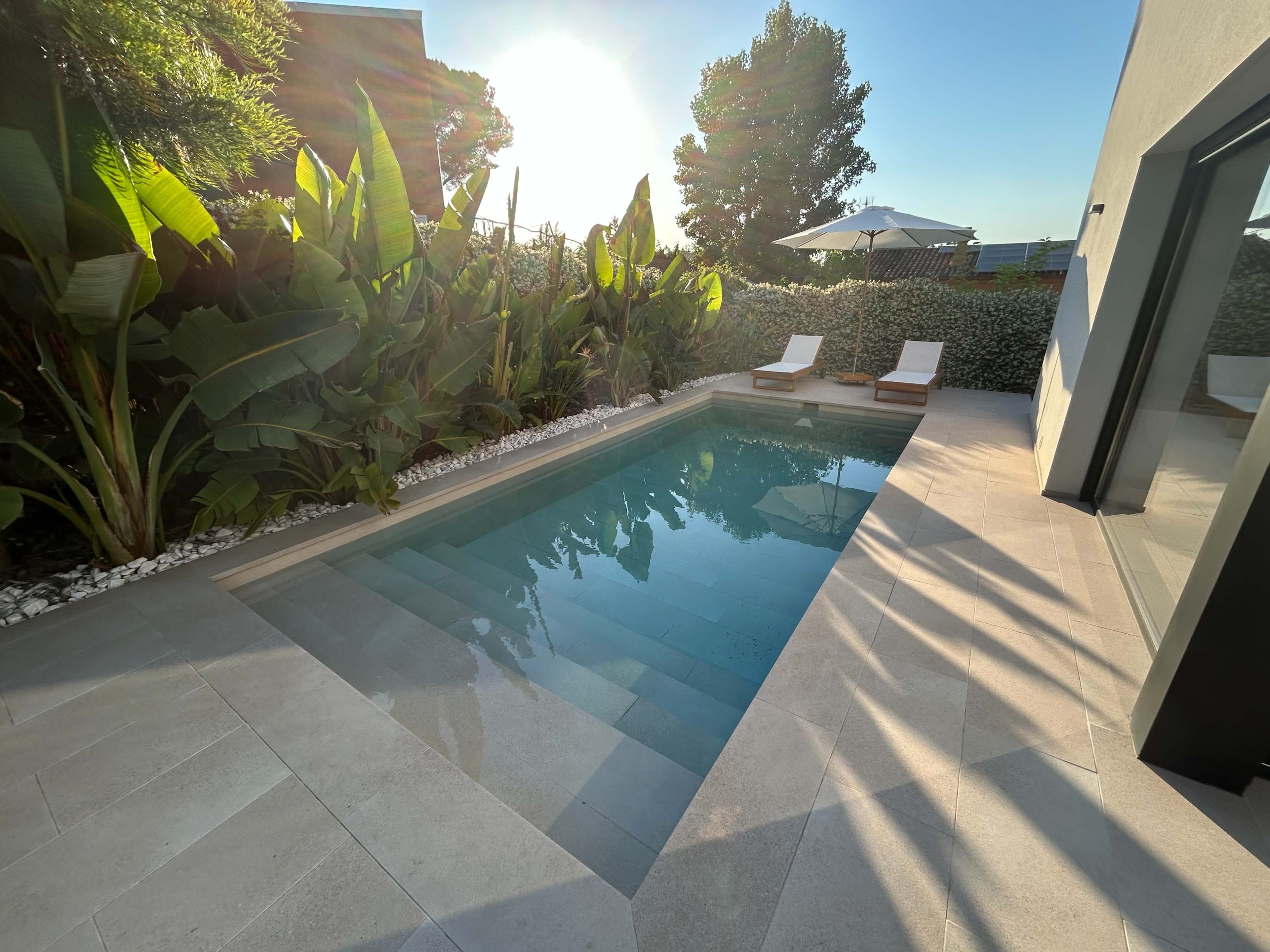 A sparkling pool is framed by tropical plants and two lounge chairs on a sunny afternoon.