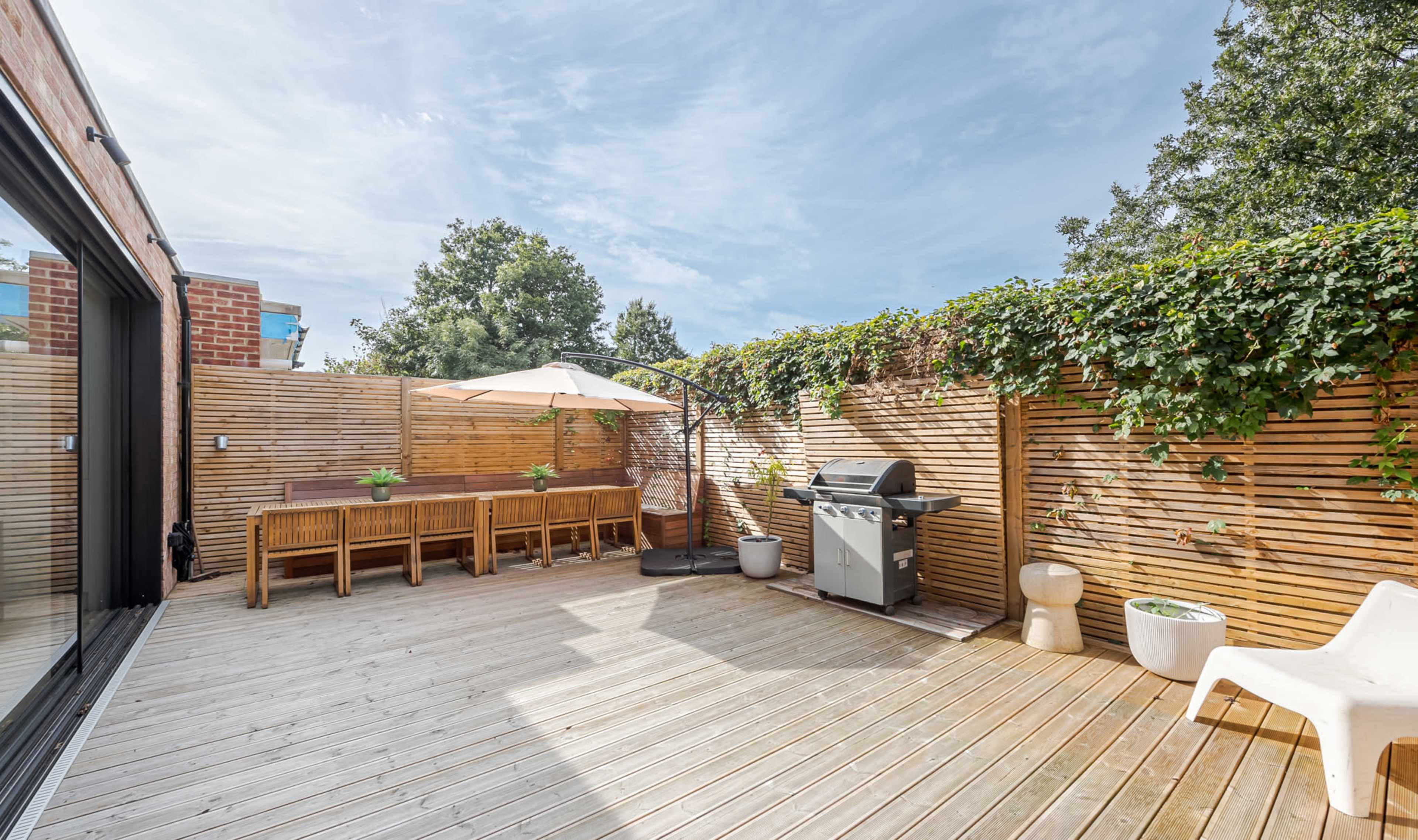 A wooden deck patio with outdoor furniture, a grill, and a parasol, surrounded by a wooden fence and greenery.