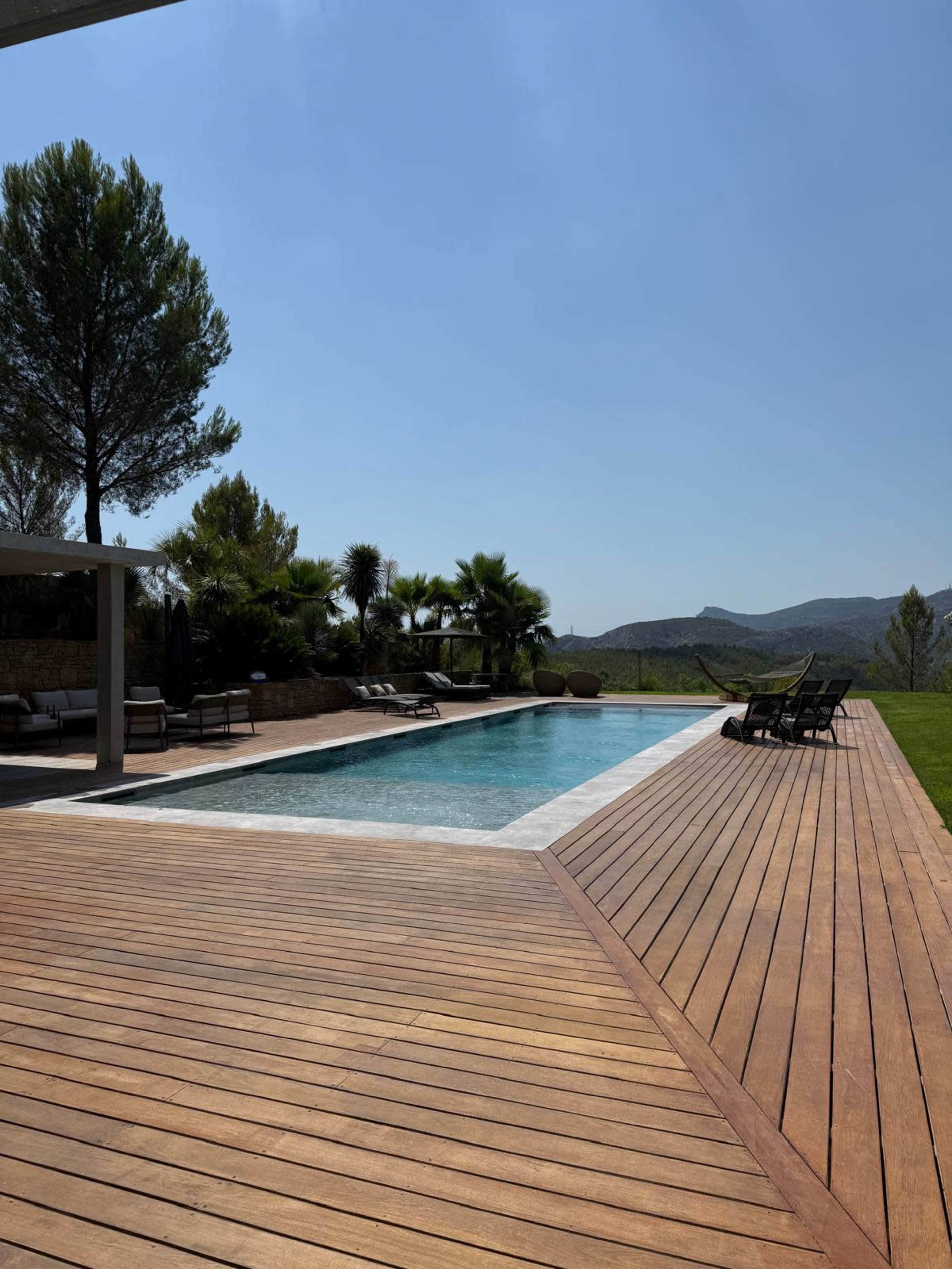 The image shows a swimming pool surrounded by wooden decking and lush greenery, with mountains visible in the background.