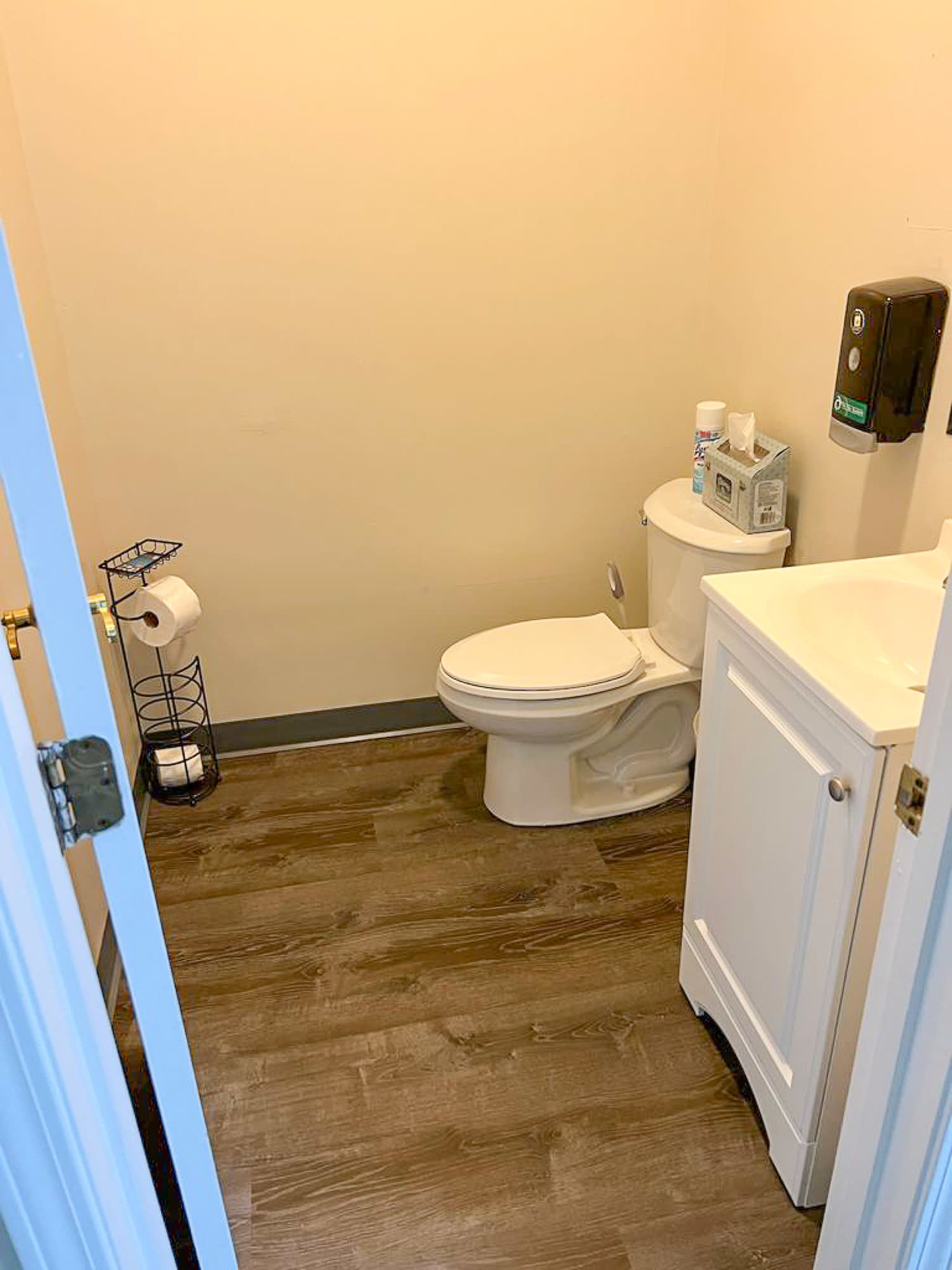 The image shows a small bathroom containing a toilet, a sink with a cabinet, a toilet paper holder, and a hand sanitizer dispenser on the wall.