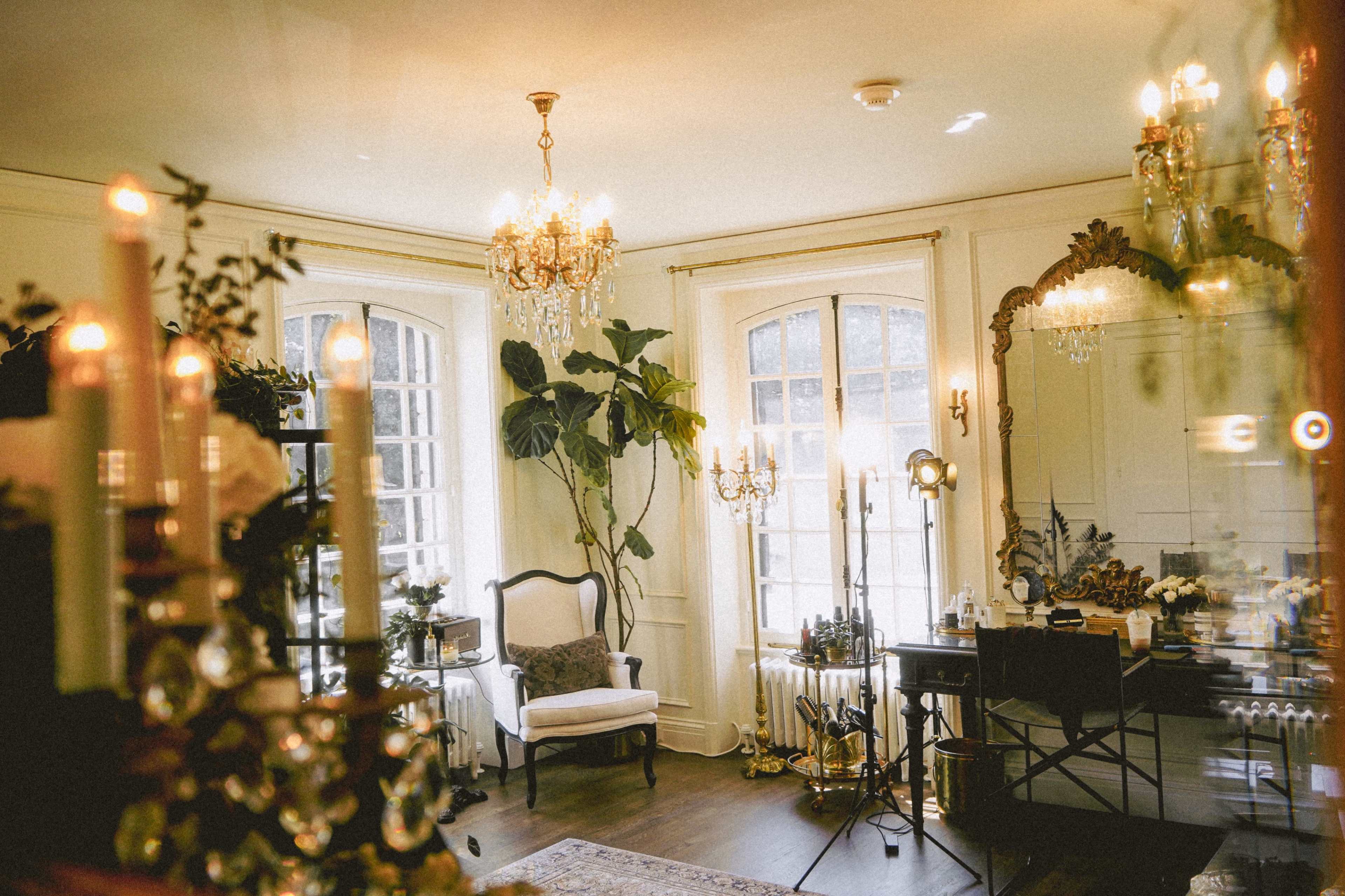 The image shows a softly lit interior of a stylish room featuring elegant chandeliers, decorative plants, and antique furnishings.