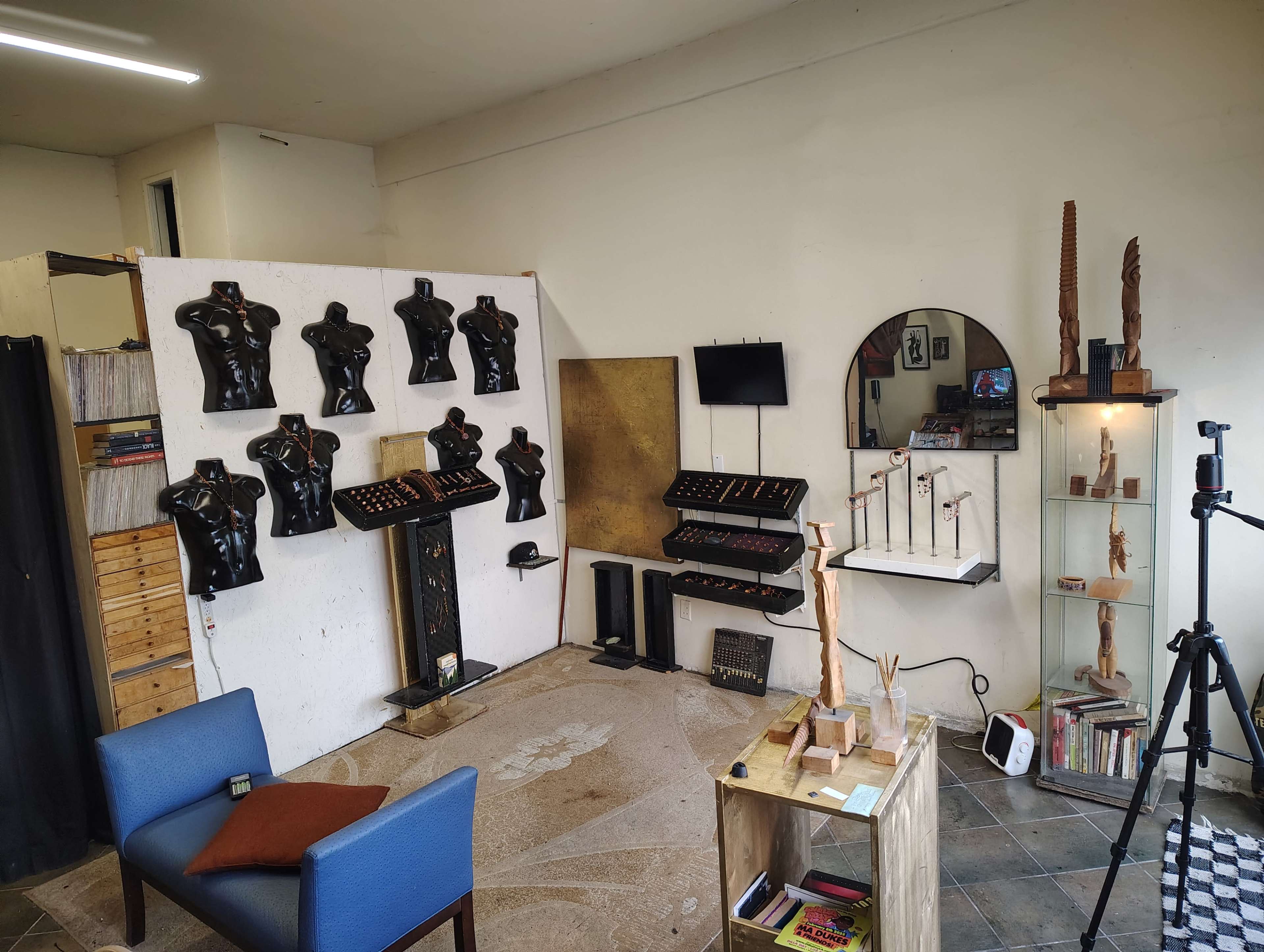 The image shows a room with a display of black torso sculptures mounted on a white wall, shelves with various items, a circular mirror, and a camera setup in the corner.