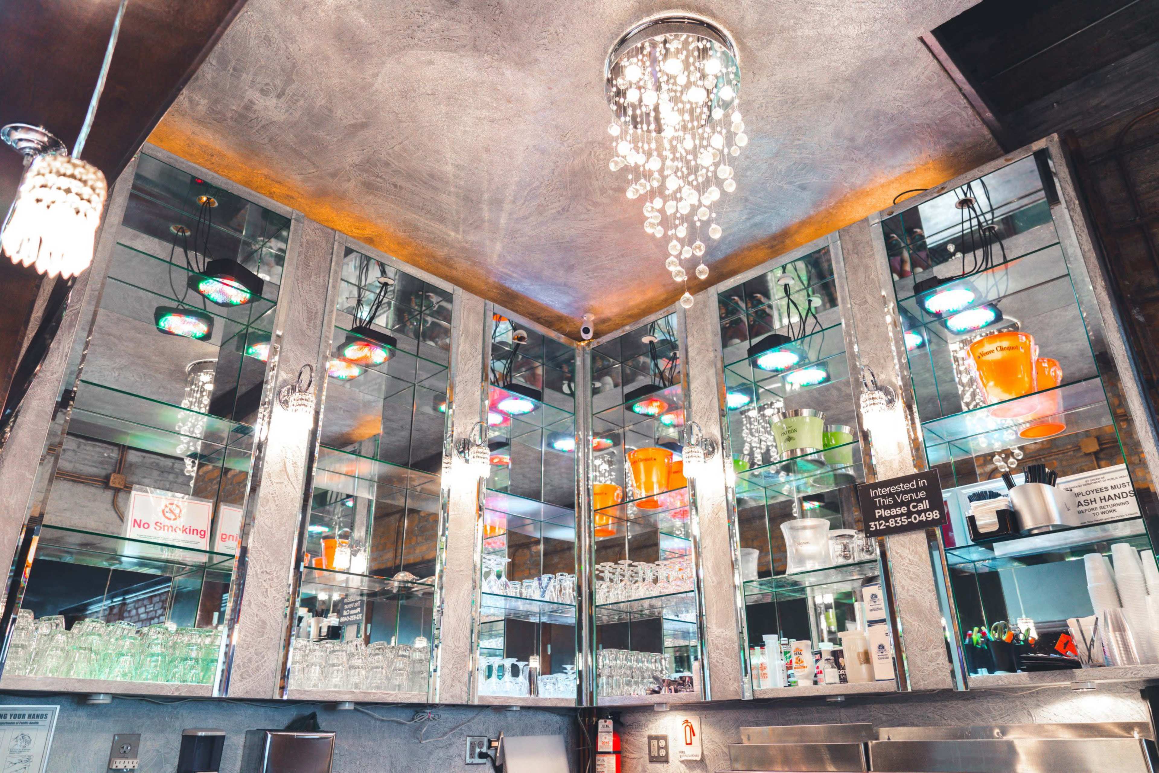 The image shows a corner view of a bar with mirrored shelves displaying various glassware and colorful drinks under decorative lighting.