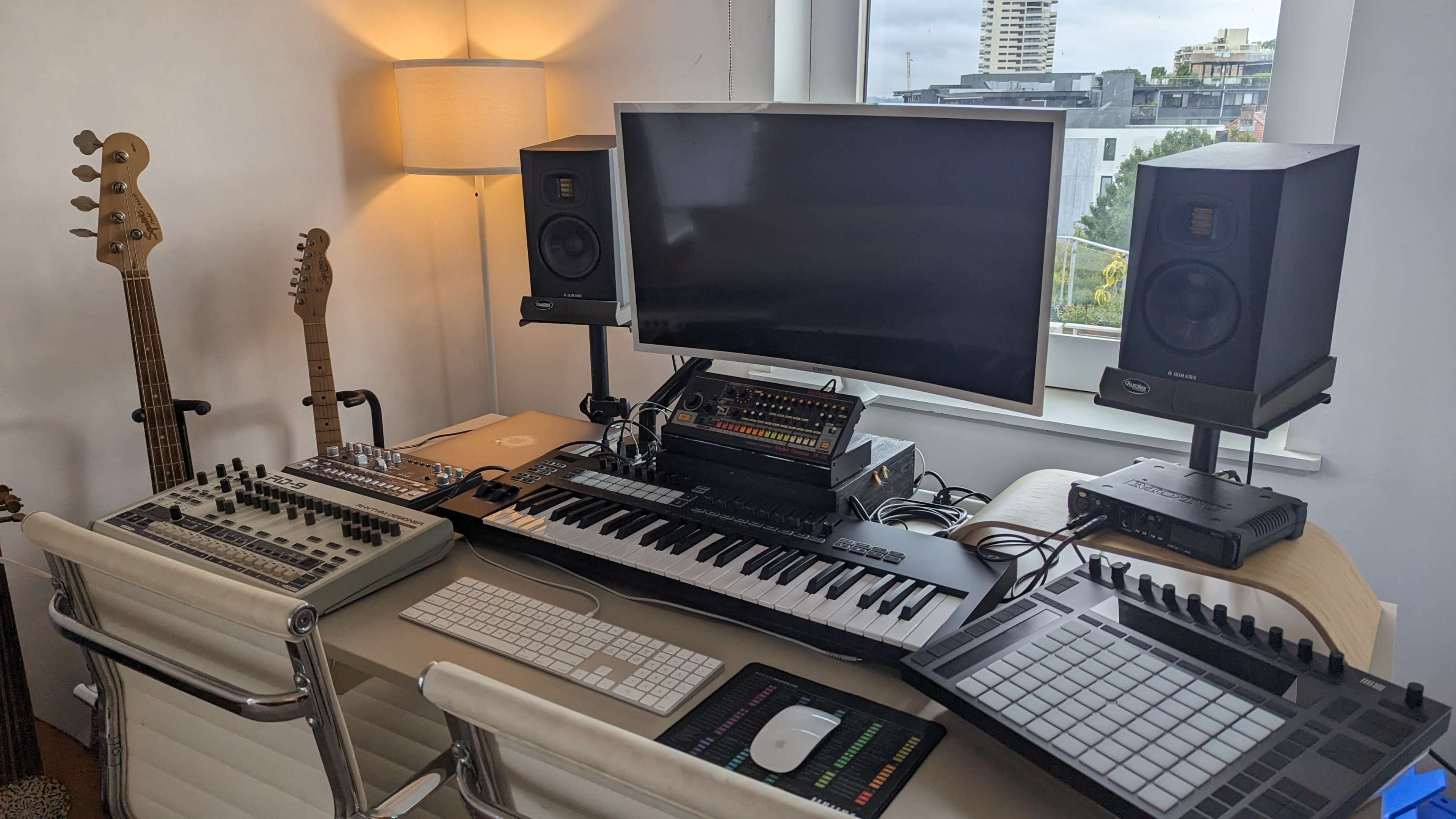A modern music production workspace featuring a computer monitor, audio speakers, a keyboard, and various electronic instruments, with guitars positioned in the background.