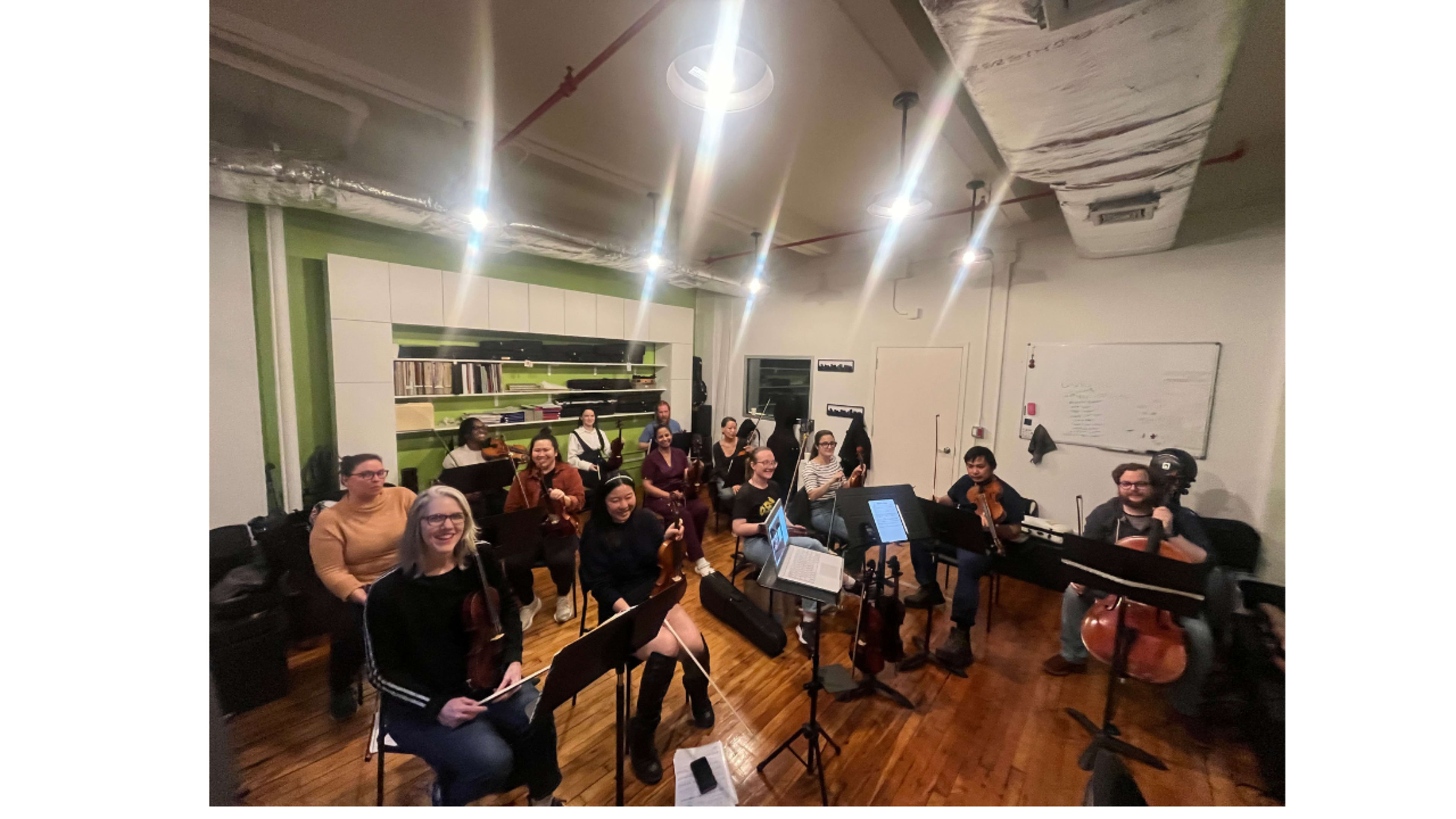 A group of musicians is seated in a room, playing string instruments during a rehearsal session.