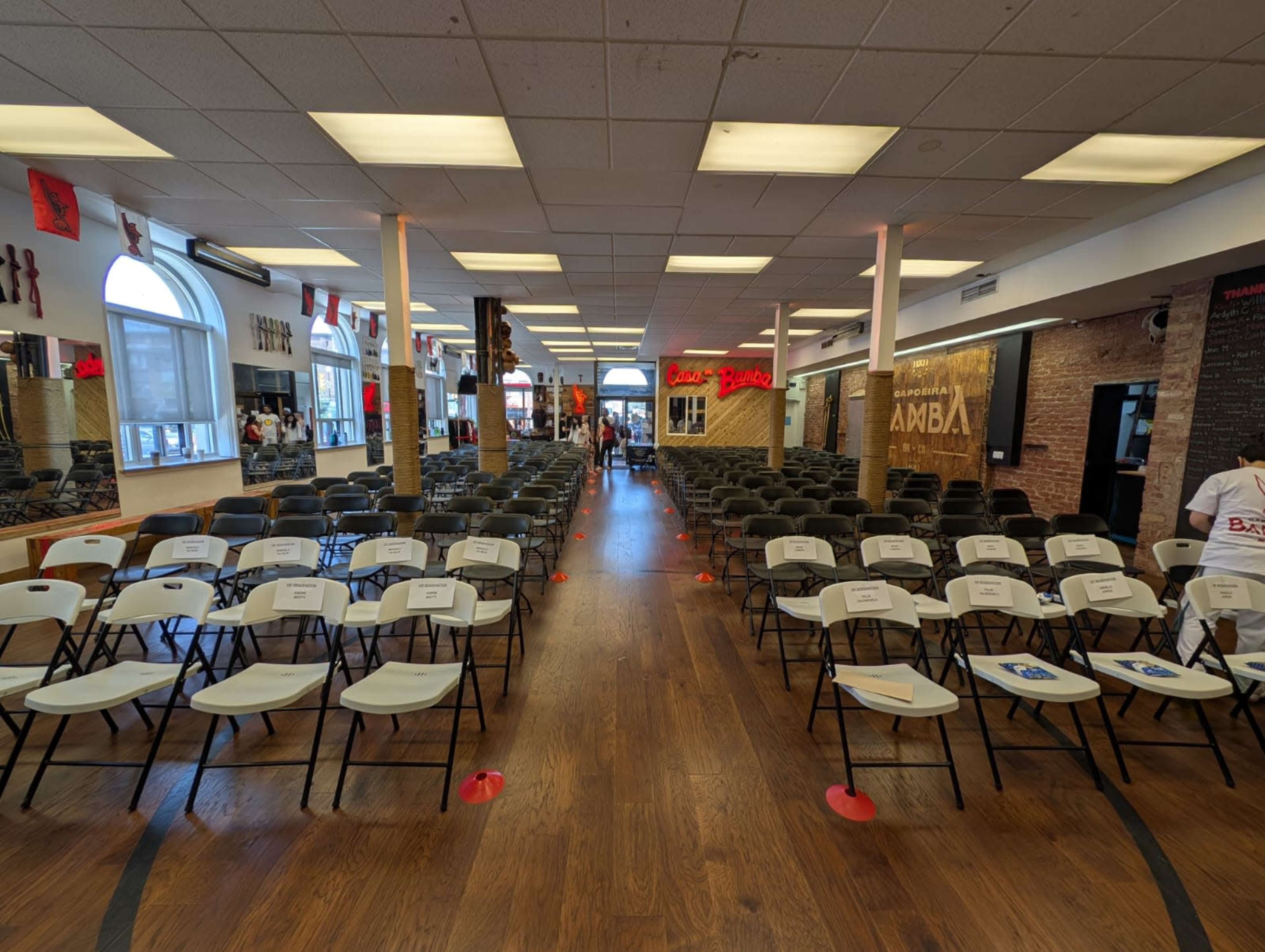 The image shows a spacious room set up with rows of white folding chairs arranged for an event, with signage and decor on the walls.