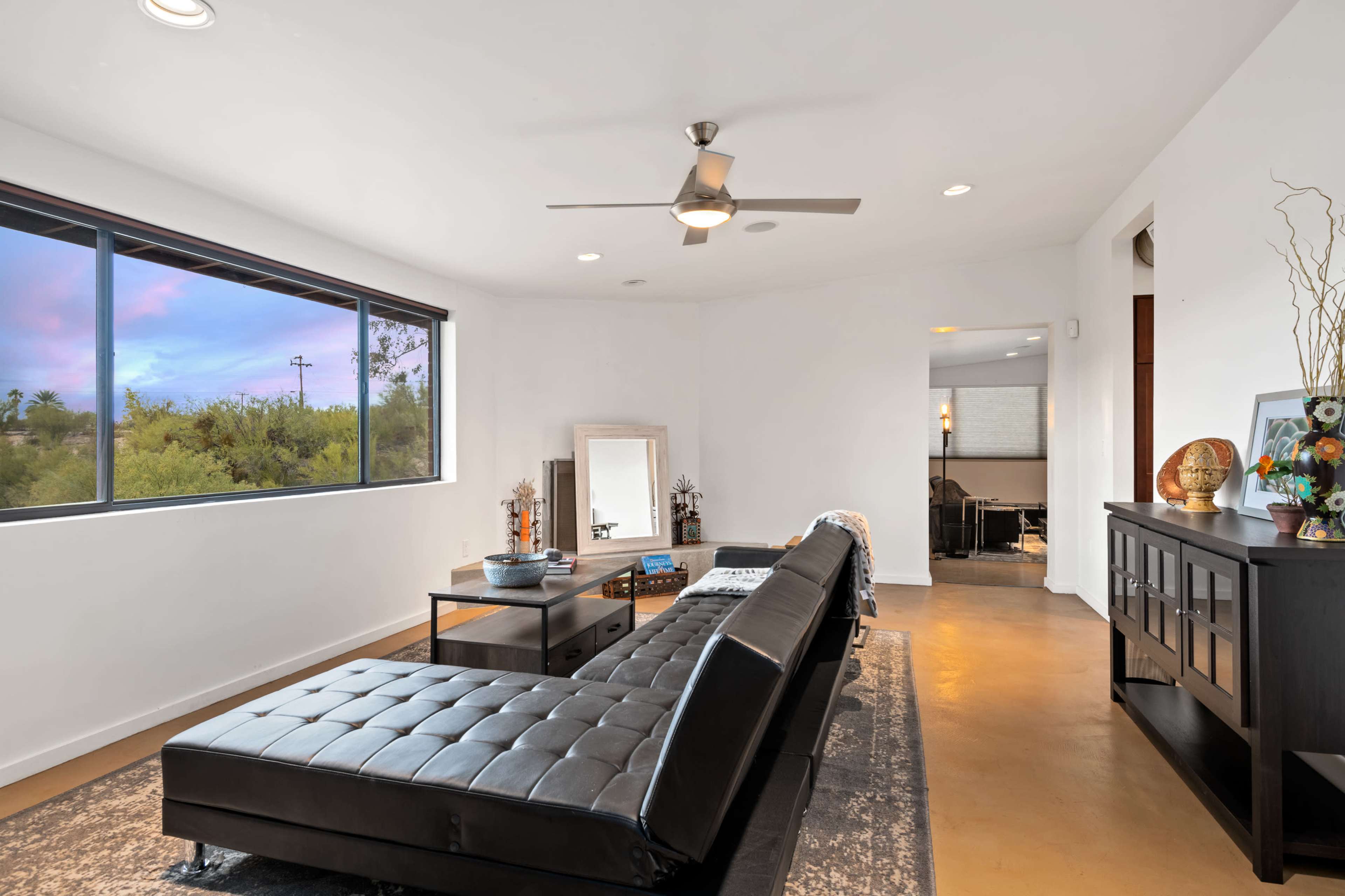 A modern living room featuring a black leather sofa, a coffee table, and large windows with a view of greenery outside.