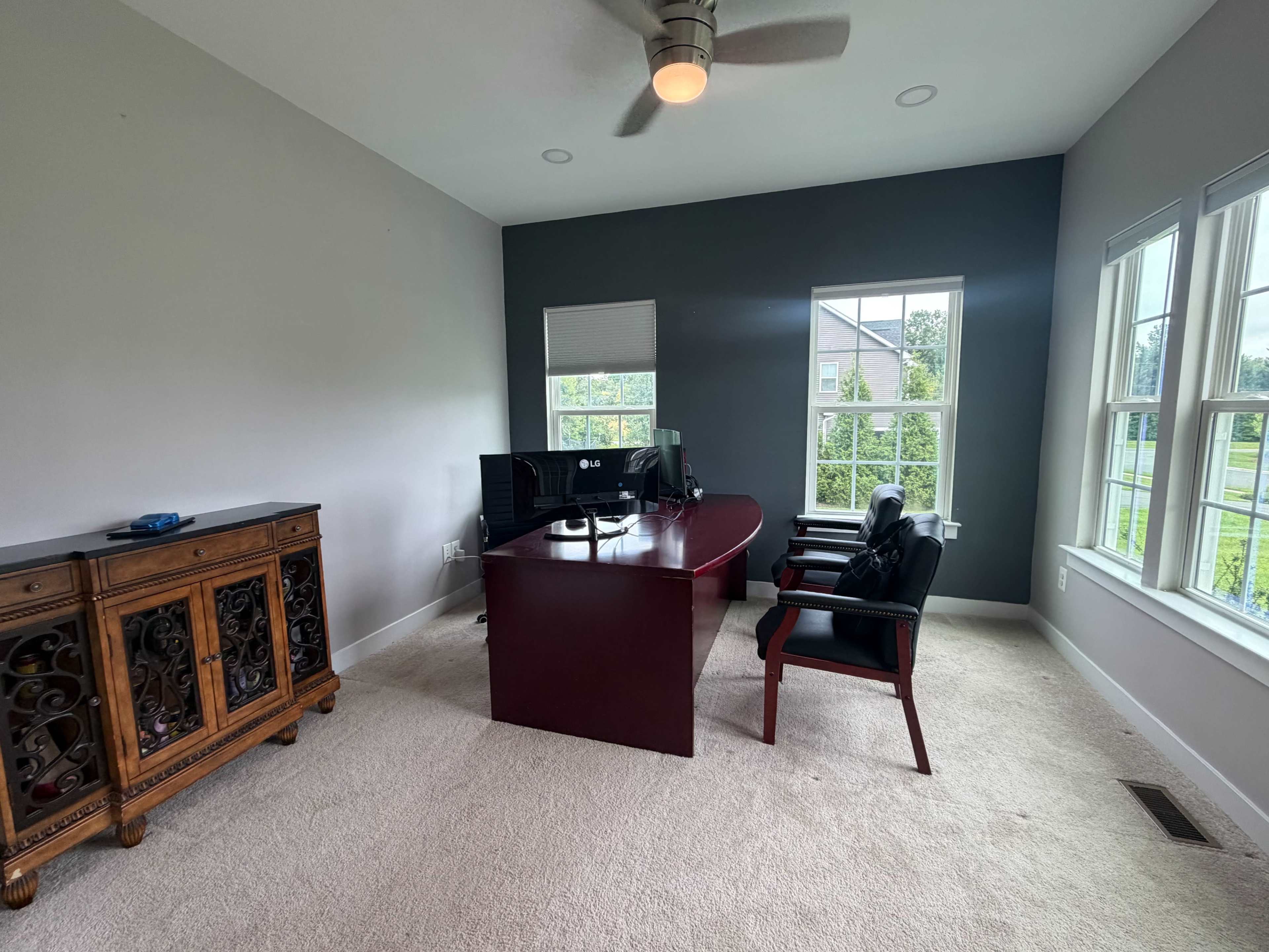The image shows a well-lit office space featuring a wooden desk, an ergonomic chair, and a vintage cabinet, with large windows providing natural light.
