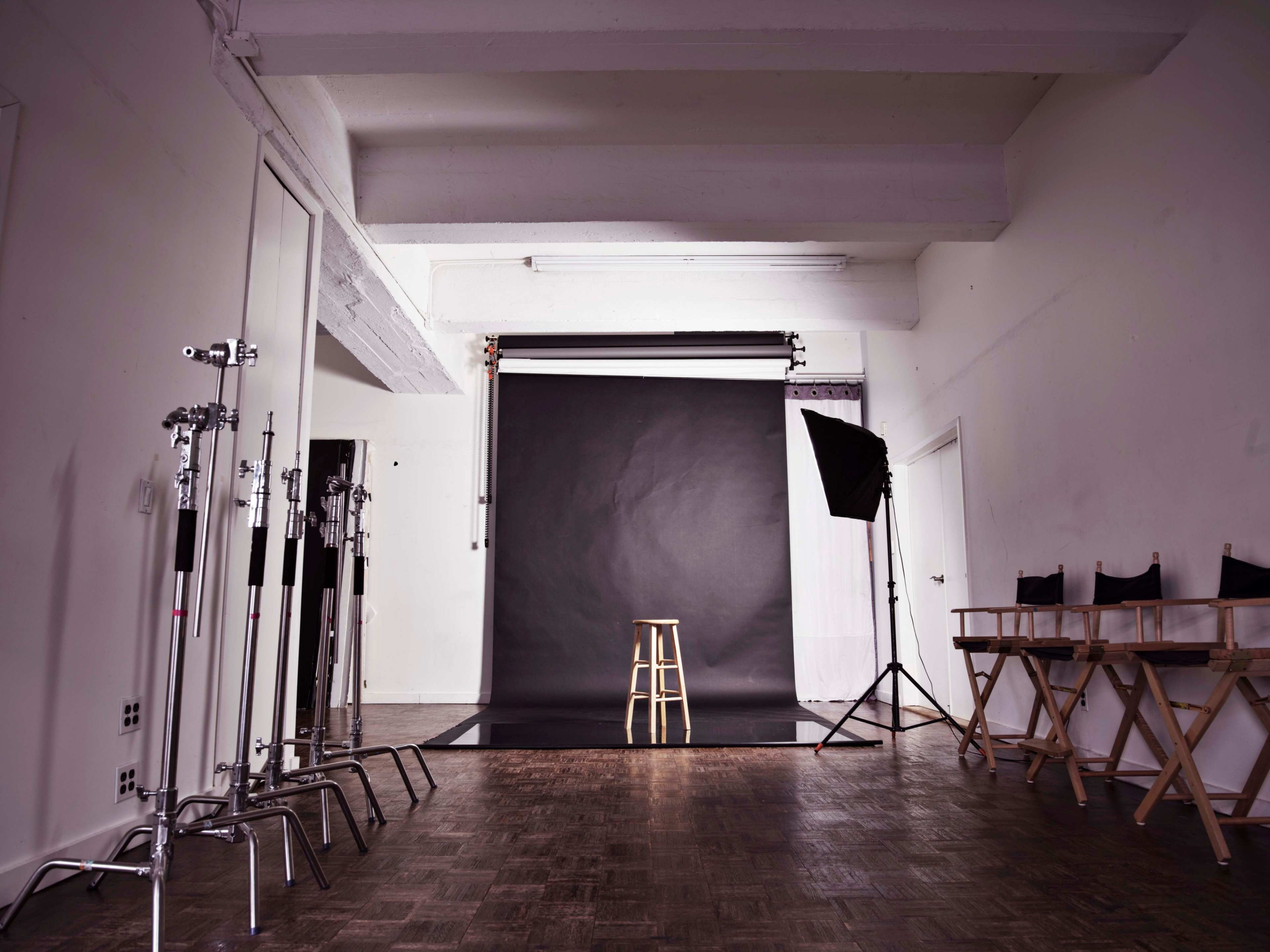 A photography studio features a black backdrop, a wooden stool in the center, and various lighting equipment along the sides.