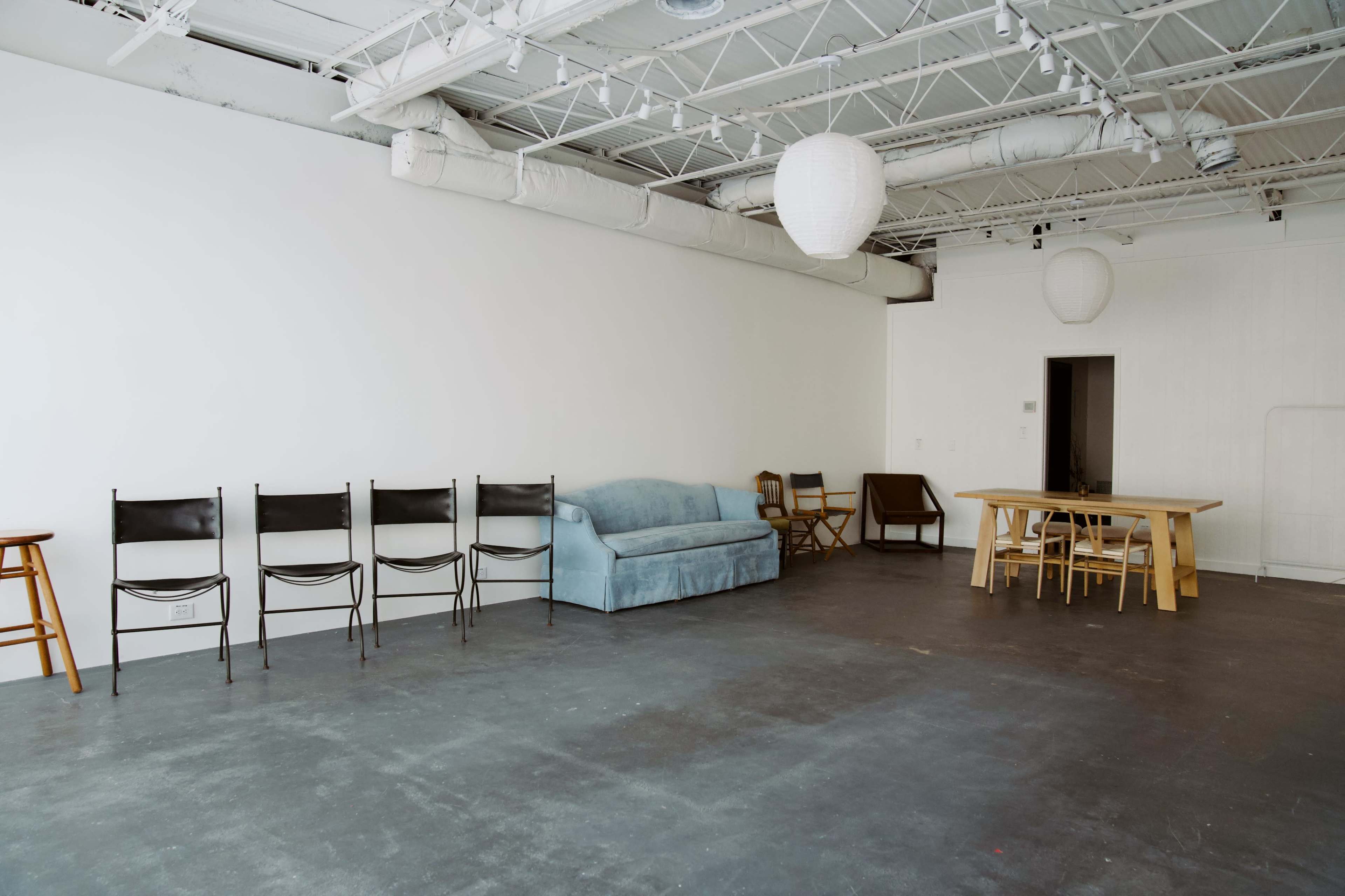 The image shows a spacious, minimalistic room furnished with a blue couch, a wooden table, and several black chairs arranged along one side.