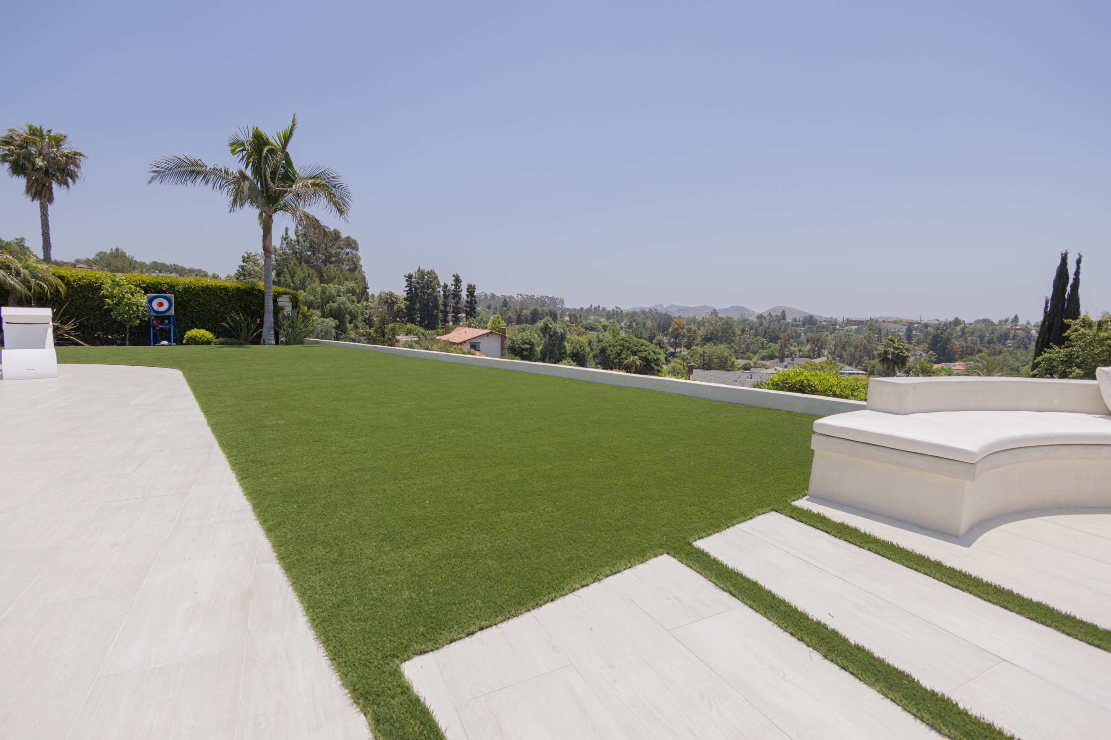 A spacious, well-maintained backyard with artificial grass, tiled pathways, and a clear view of distant trees and hills.