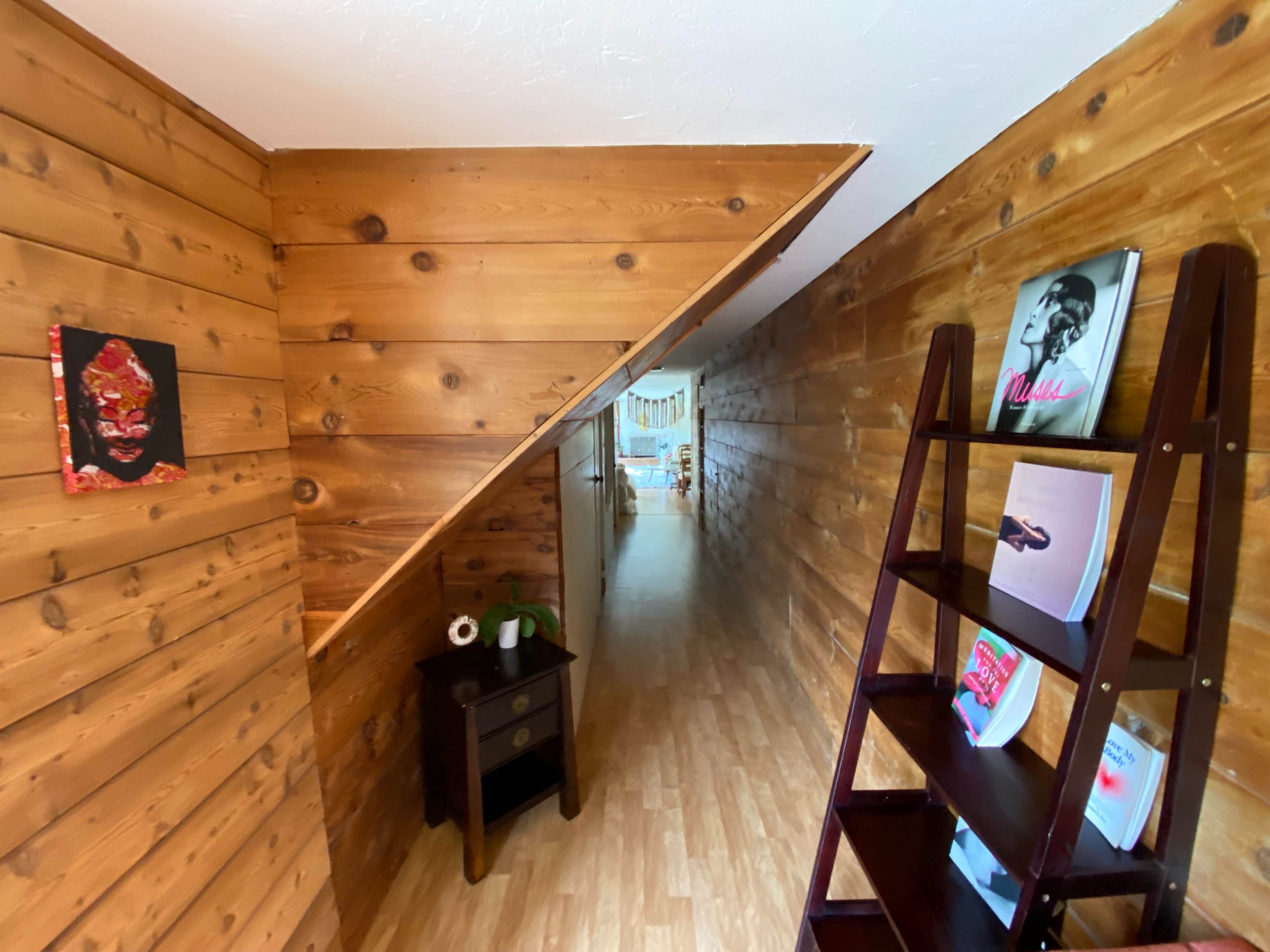 A wooden-paneled hallway leading to a brightly lit room, with a shelf displaying magazines on one side and wall art on the opposite side.