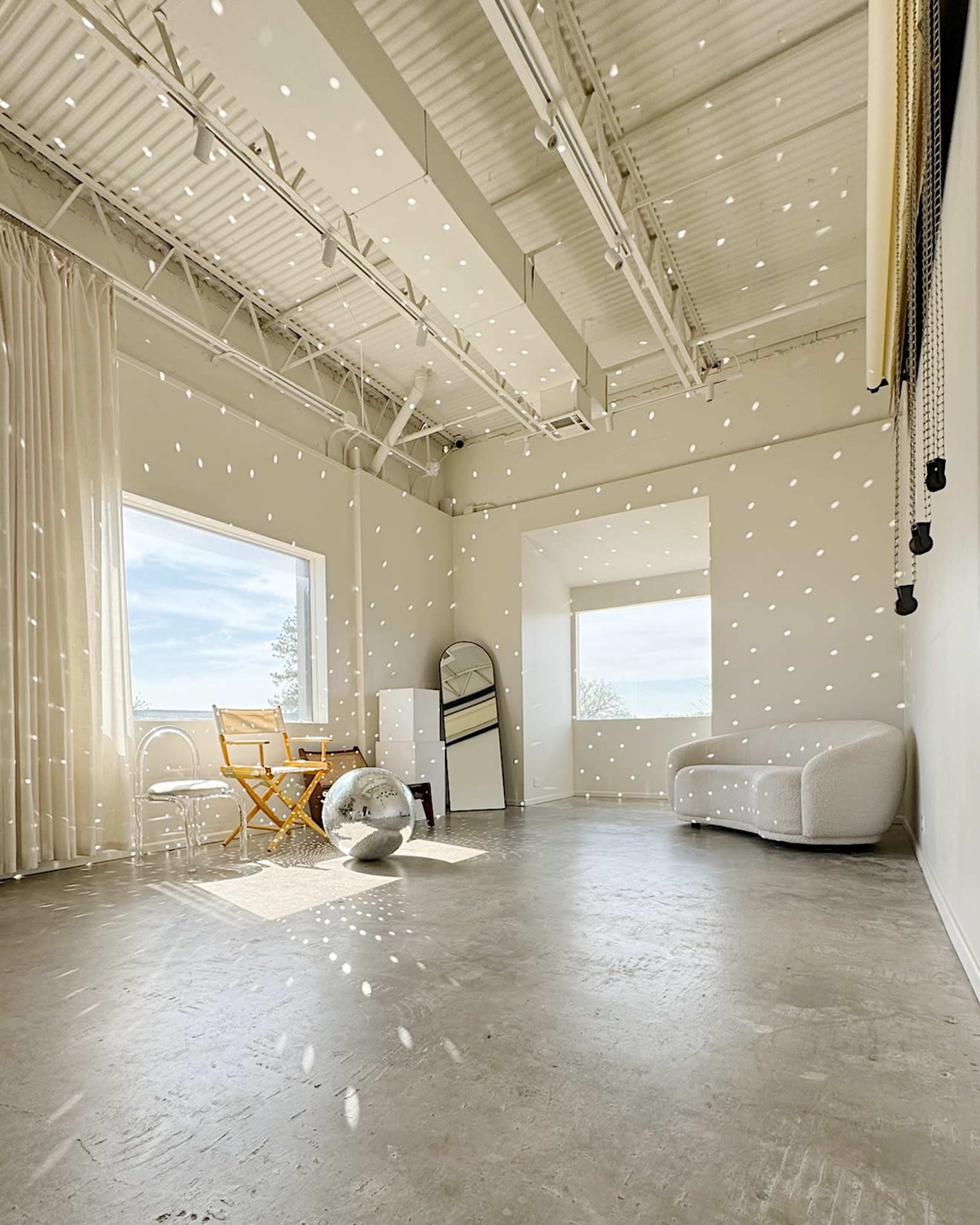 A bright, spacious room features large windows, minimalist furniture, and a reflective disco ball on the floor.