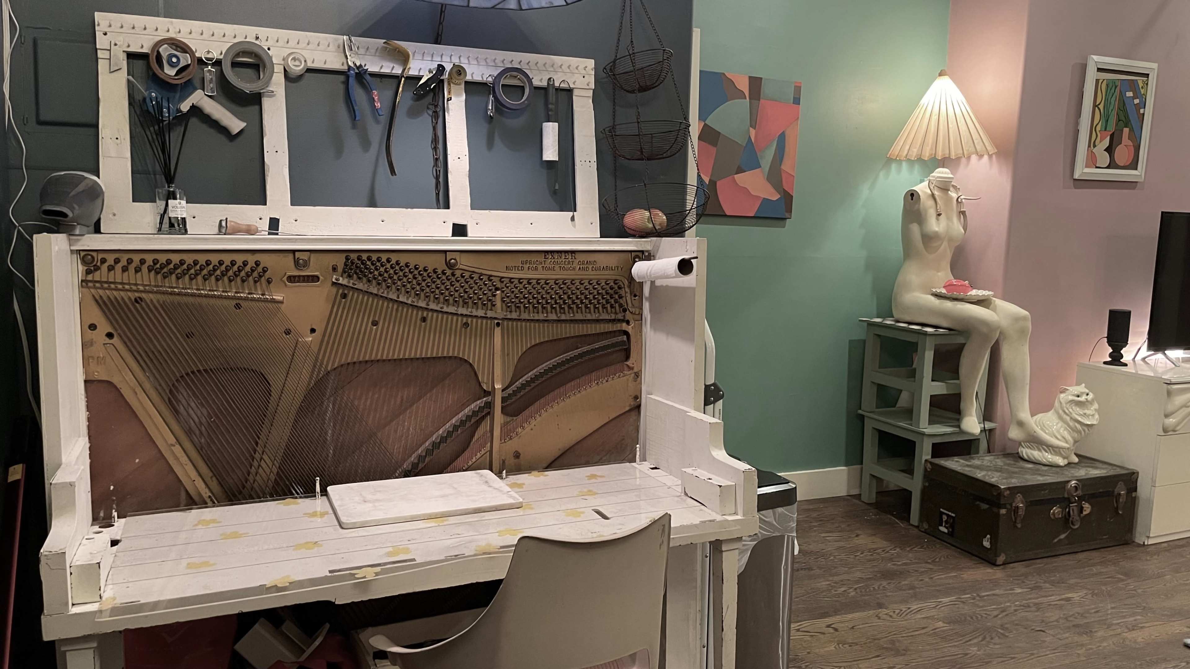 The image shows a creatively designed workspace featuring an upright piano's interior as a backdrop, surrounded by various tools and a sculpture on a nearby pedestal.
