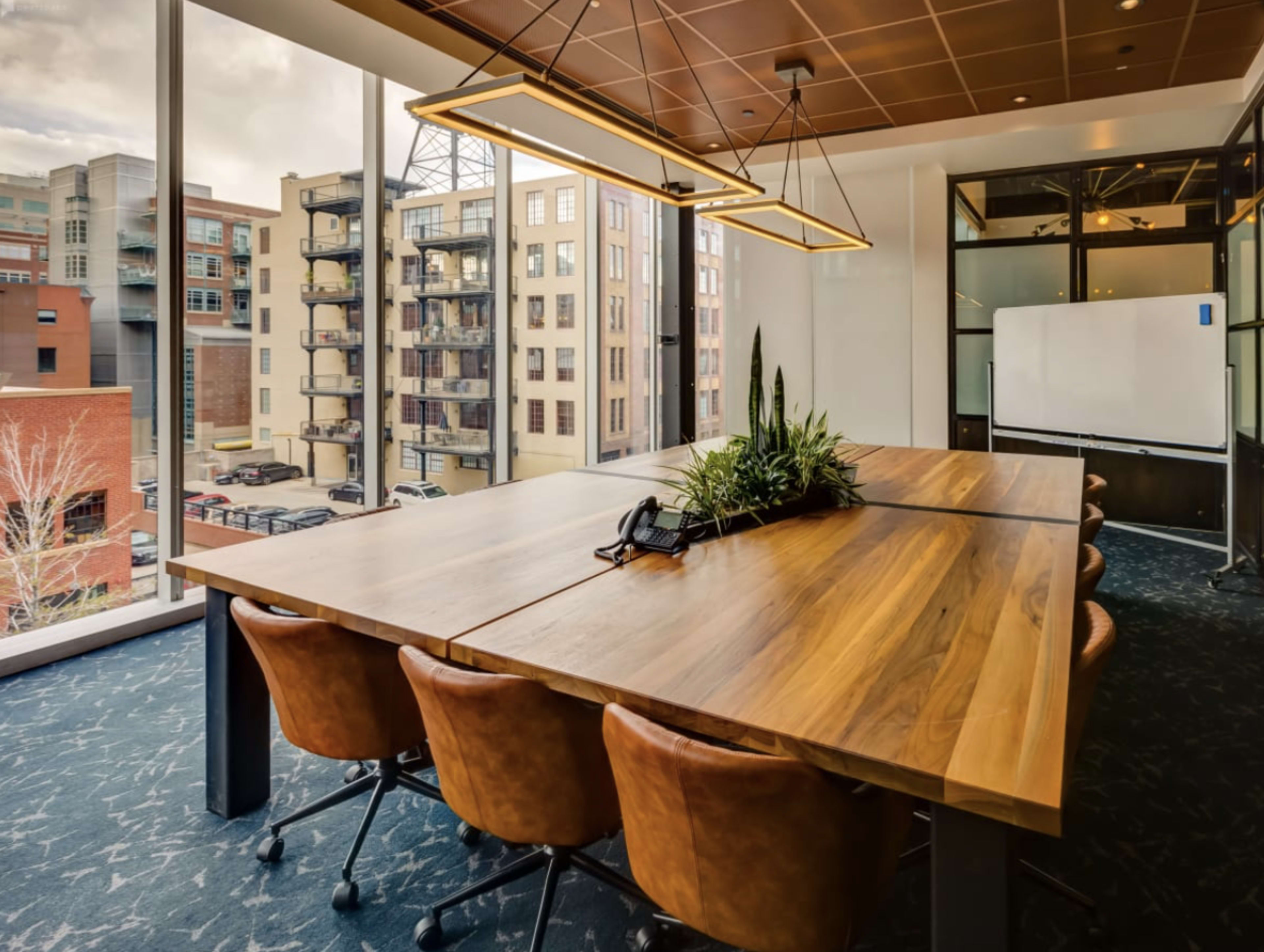 A large wooden conference table with brown chairs is situated in a well-lit office with large windows showing a view of neighboring buildings.
