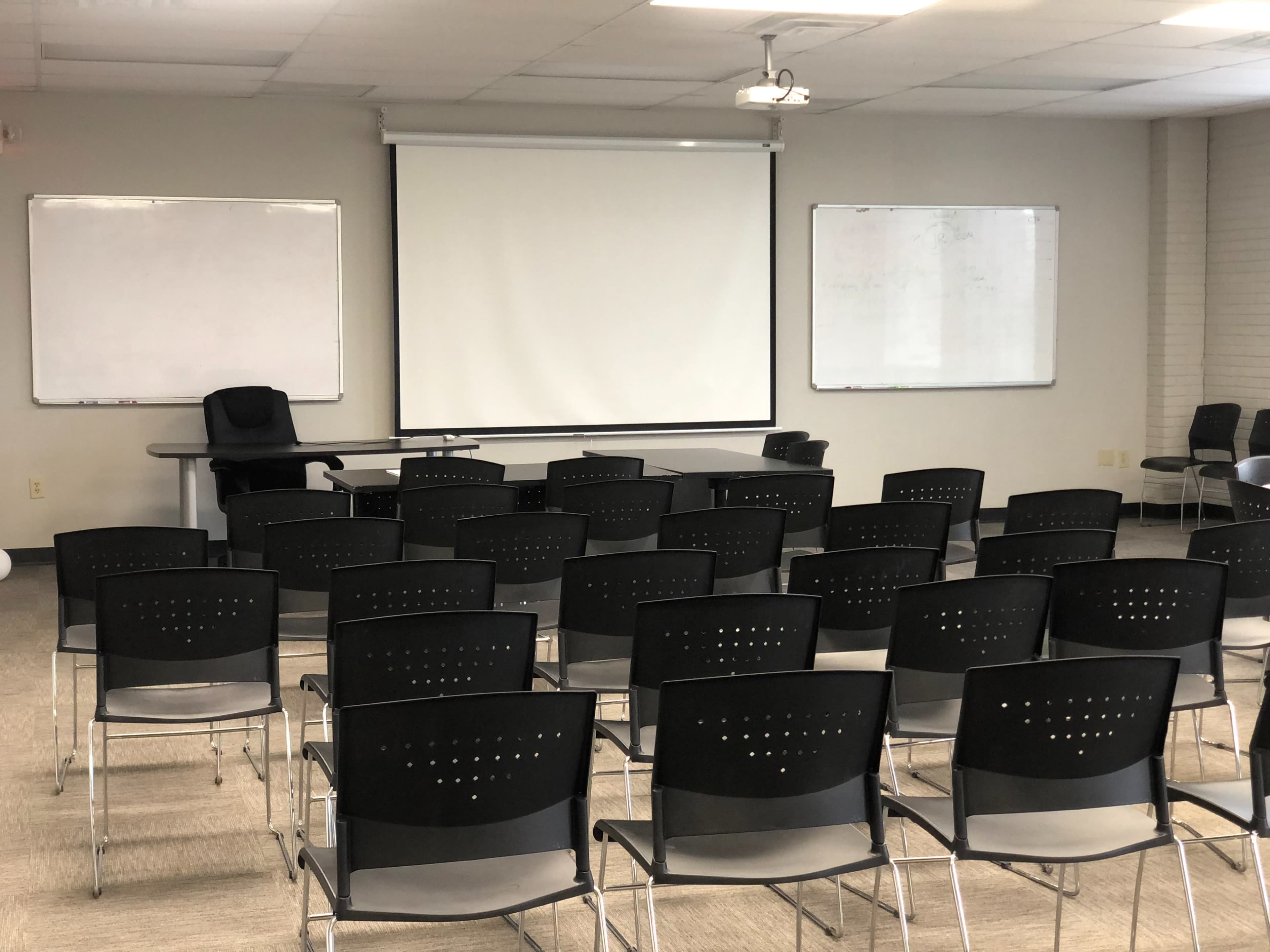 A classroom setup with rows of black chairs facing a large projection screen, flanked by two whiteboards.