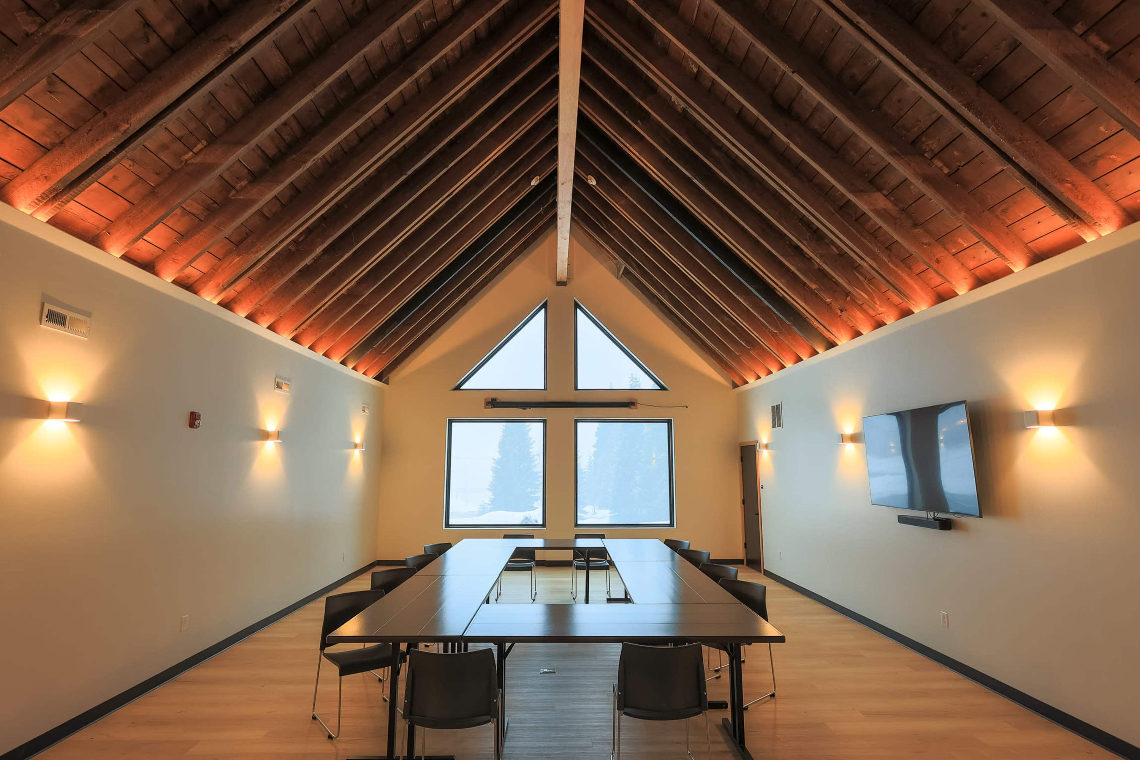 A modern meeting room features a long table surrounded by chairs under a vaulted wooden ceiling with large windows providing natural light.