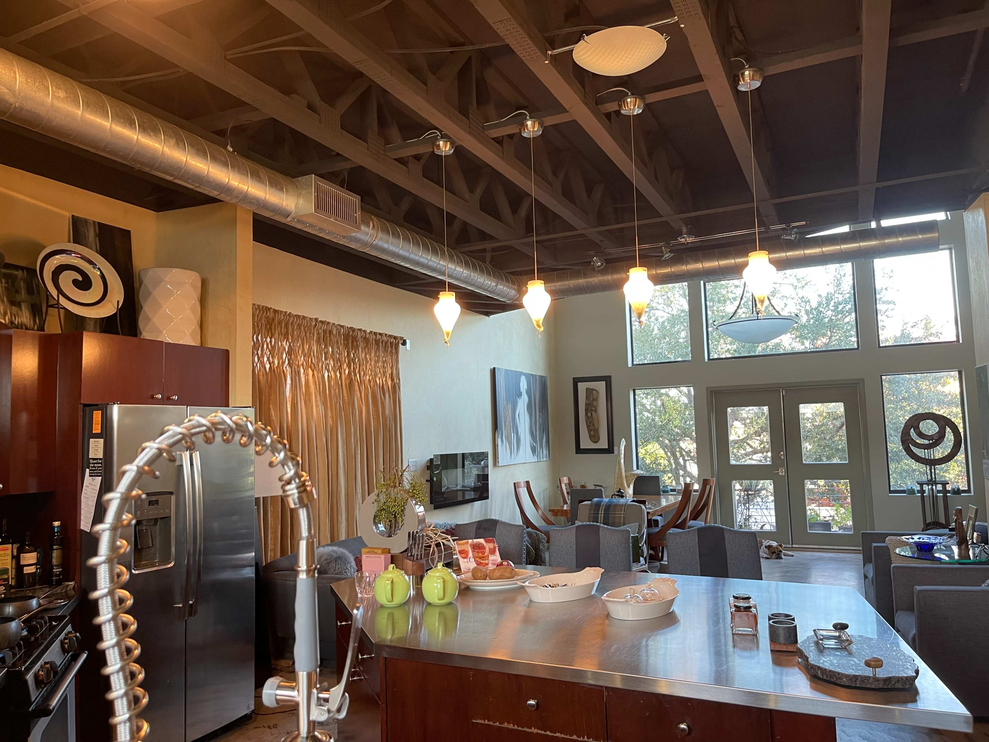 The image shows a modern kitchen with stainless steel appliances and a dining area featuring large windows that allow natural light to enter.