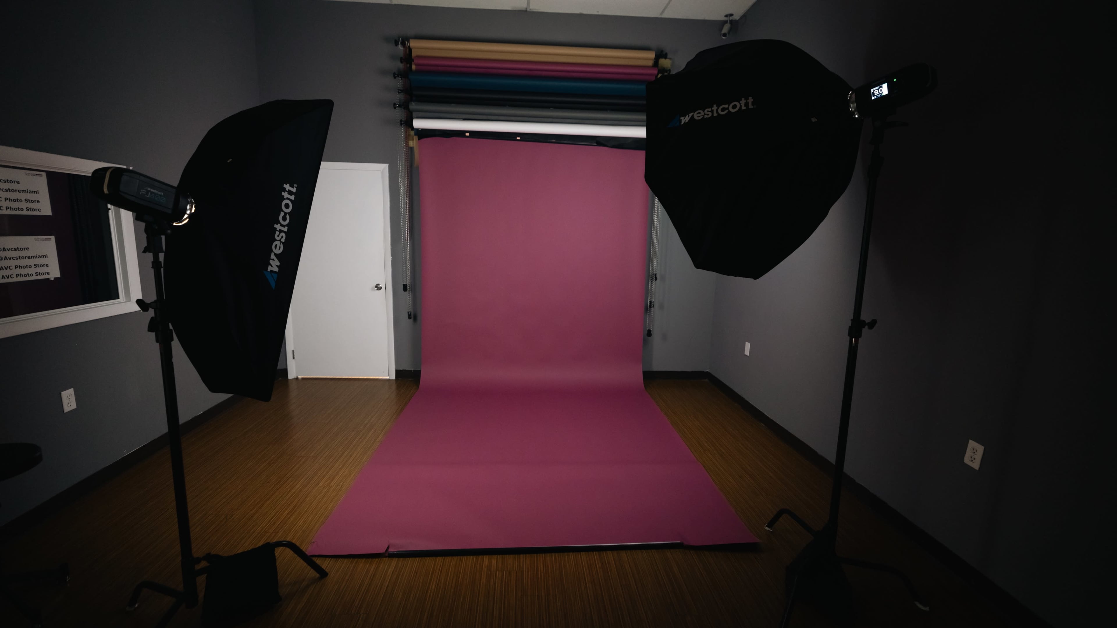 The image shows a photography studio equipped with two softbox lights and a solid pink backdrop set against a gray wall.