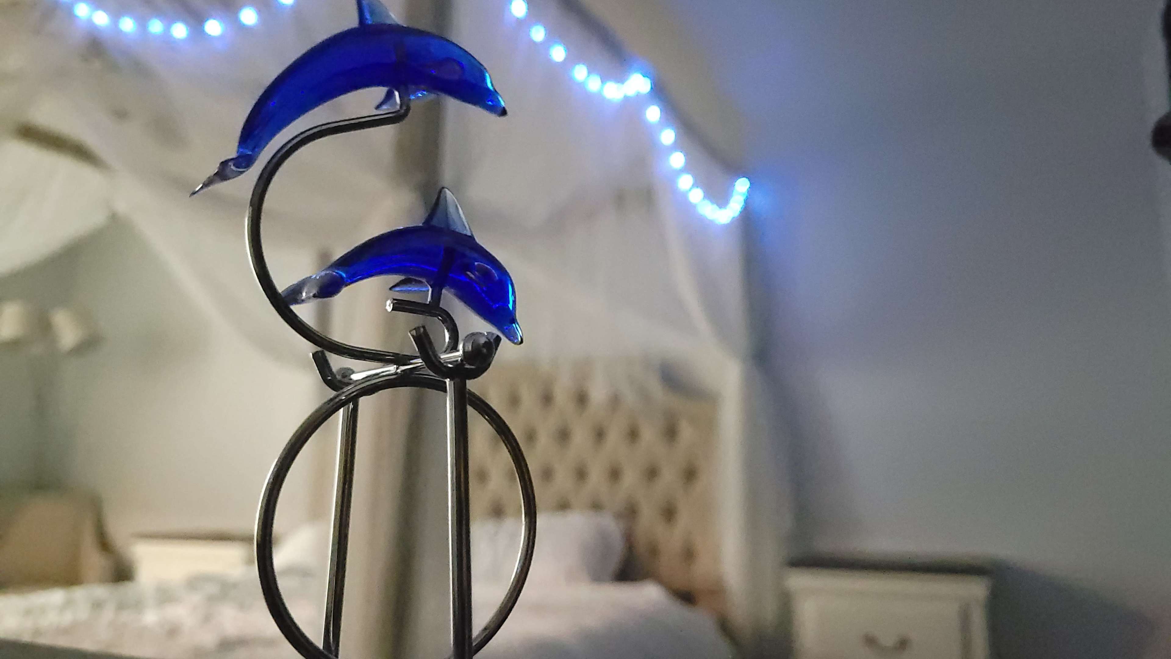 A metal holder featuring two blue dolphin figurines is placed in front of a softly lit bedroom with a bed and decorative string lights.