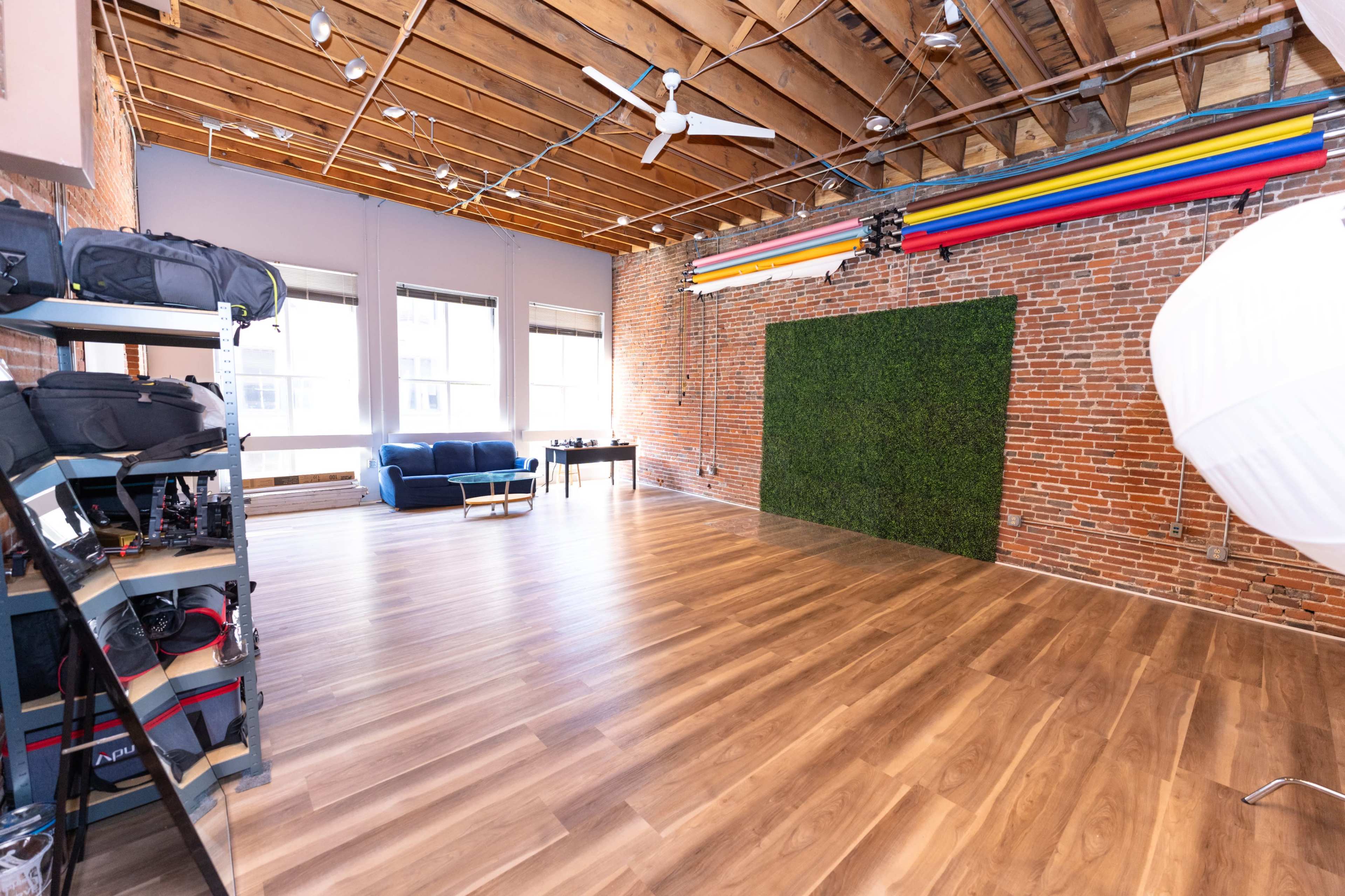 The image shows a spacious studio with wooden flooring, exposed brick walls, and a green backdrop, along with photography equipment.