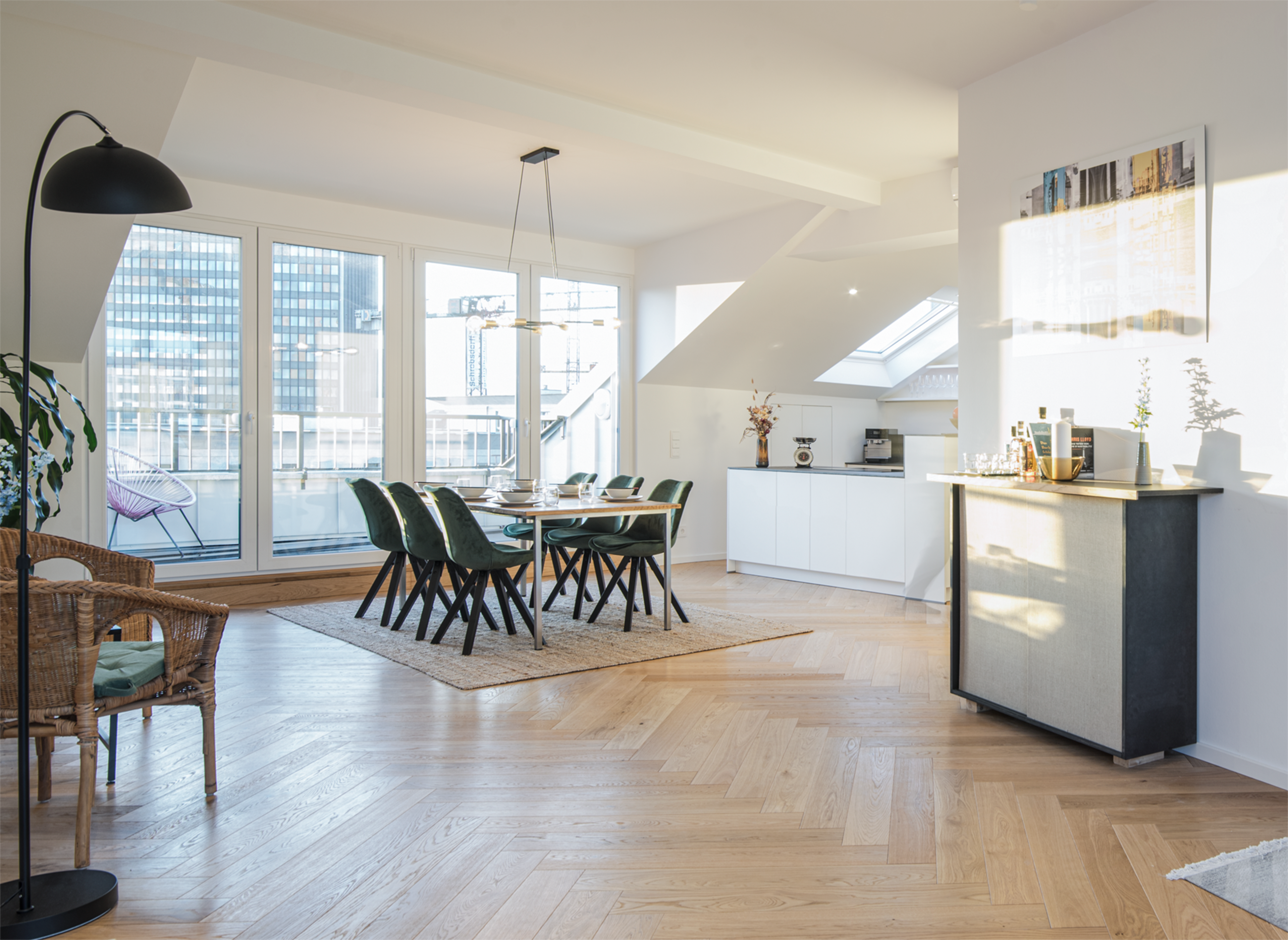 A modern dining area with a large table surrounded by green chairs, adjacent to a kitchen counter and bright windows overlooking a cityscape.