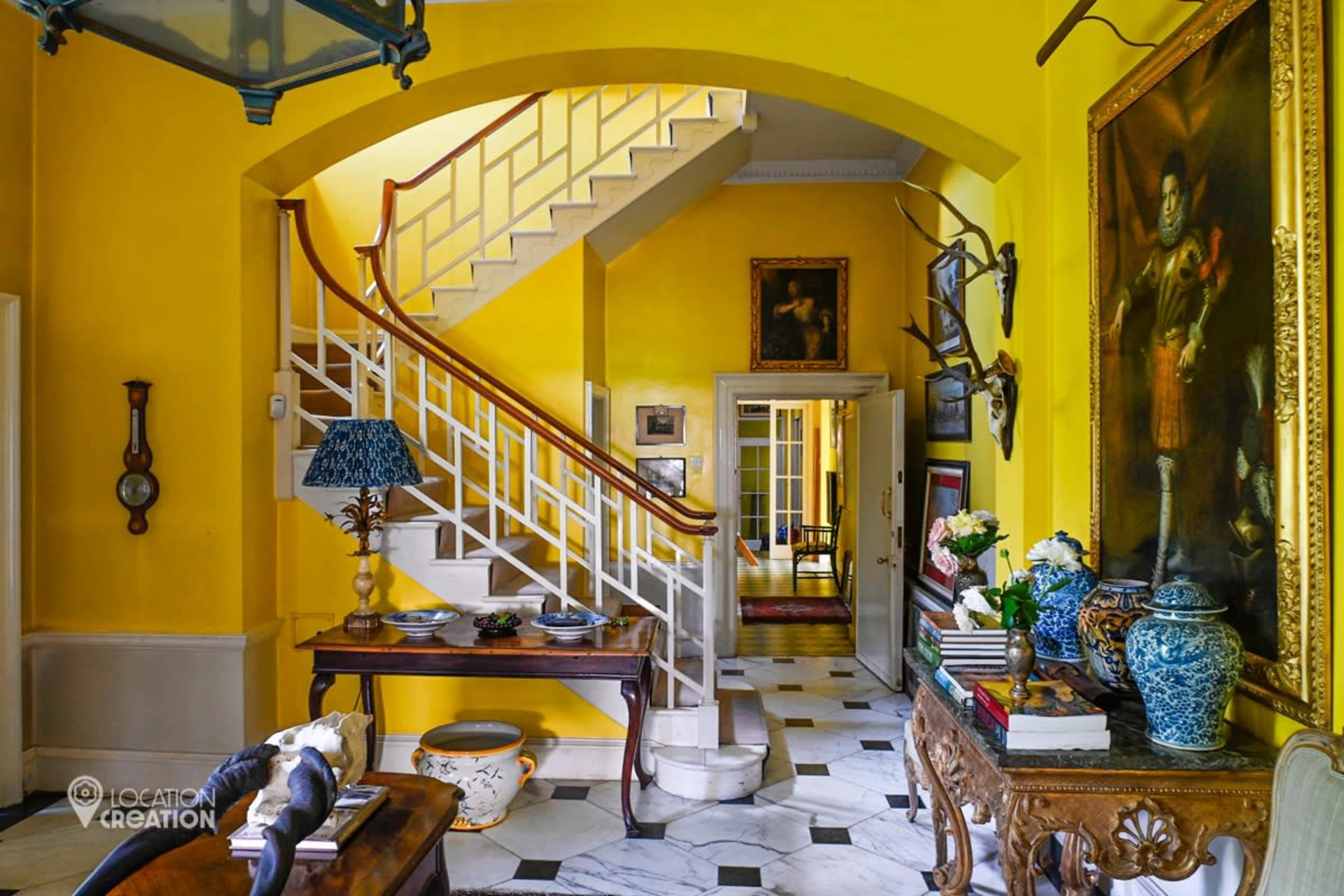 A bright yellow hallway features a staircase, decorative artwork, and furniture, with marble flooring adding to the interior design.