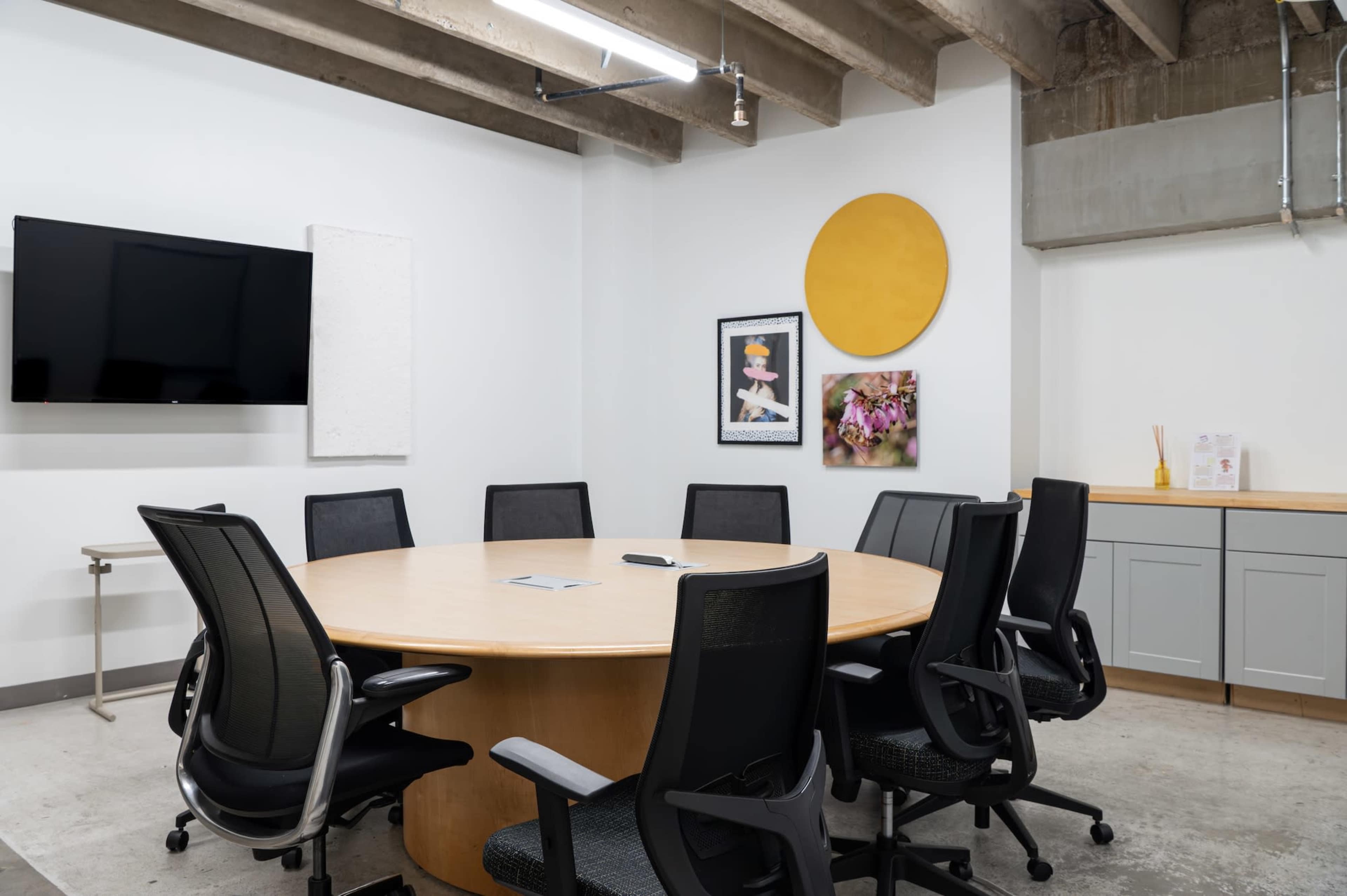 A circular wooden conference table surrounded by black ergonomic chairs is set up in a modern meeting room with a television screen and artwork on the walls.
