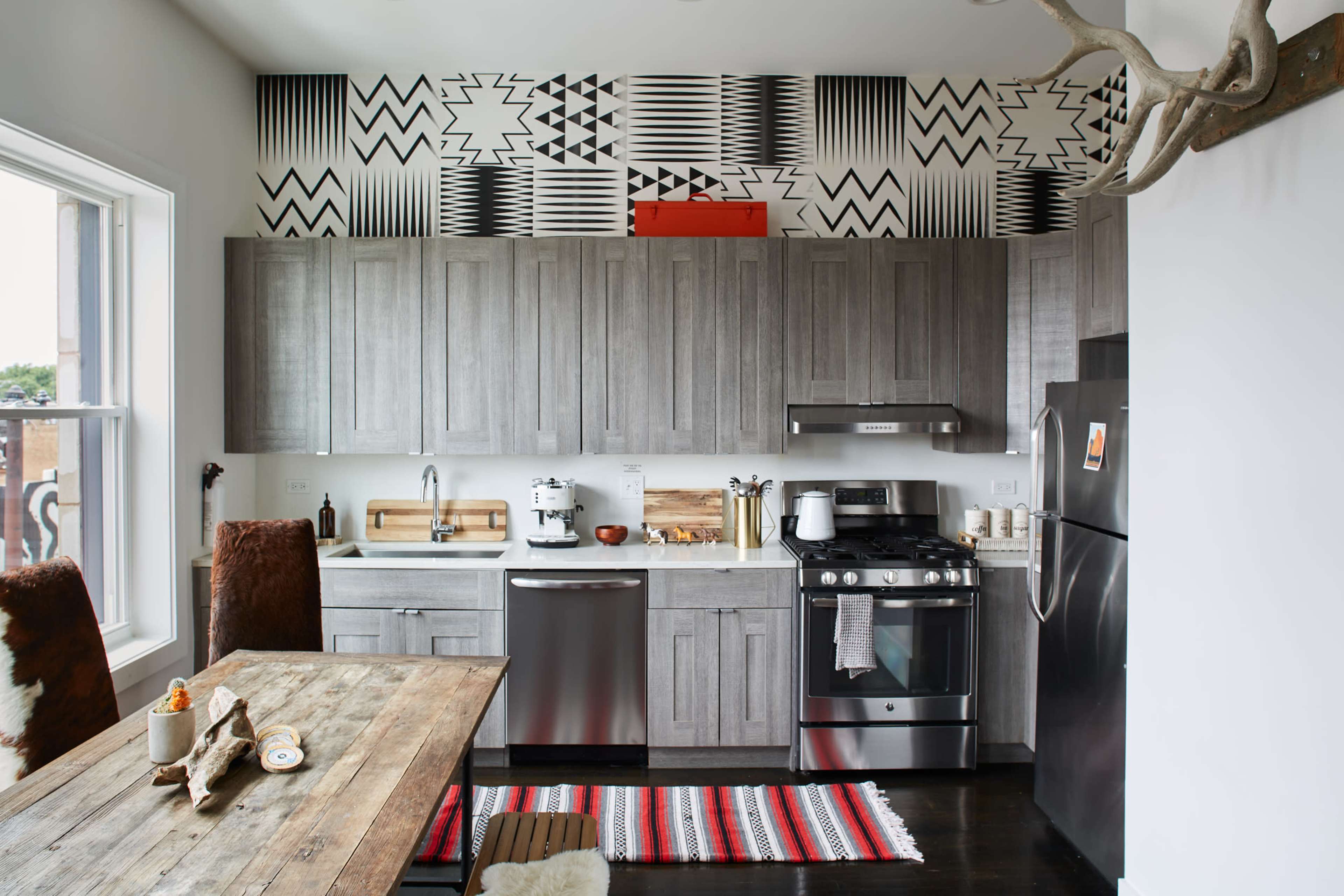 The image shows a modern kitchen with gray cabinetry, stainless steel appliances, and a patterned wallpaper above the cabinets, paired with a wooden dining table and rustic decor.