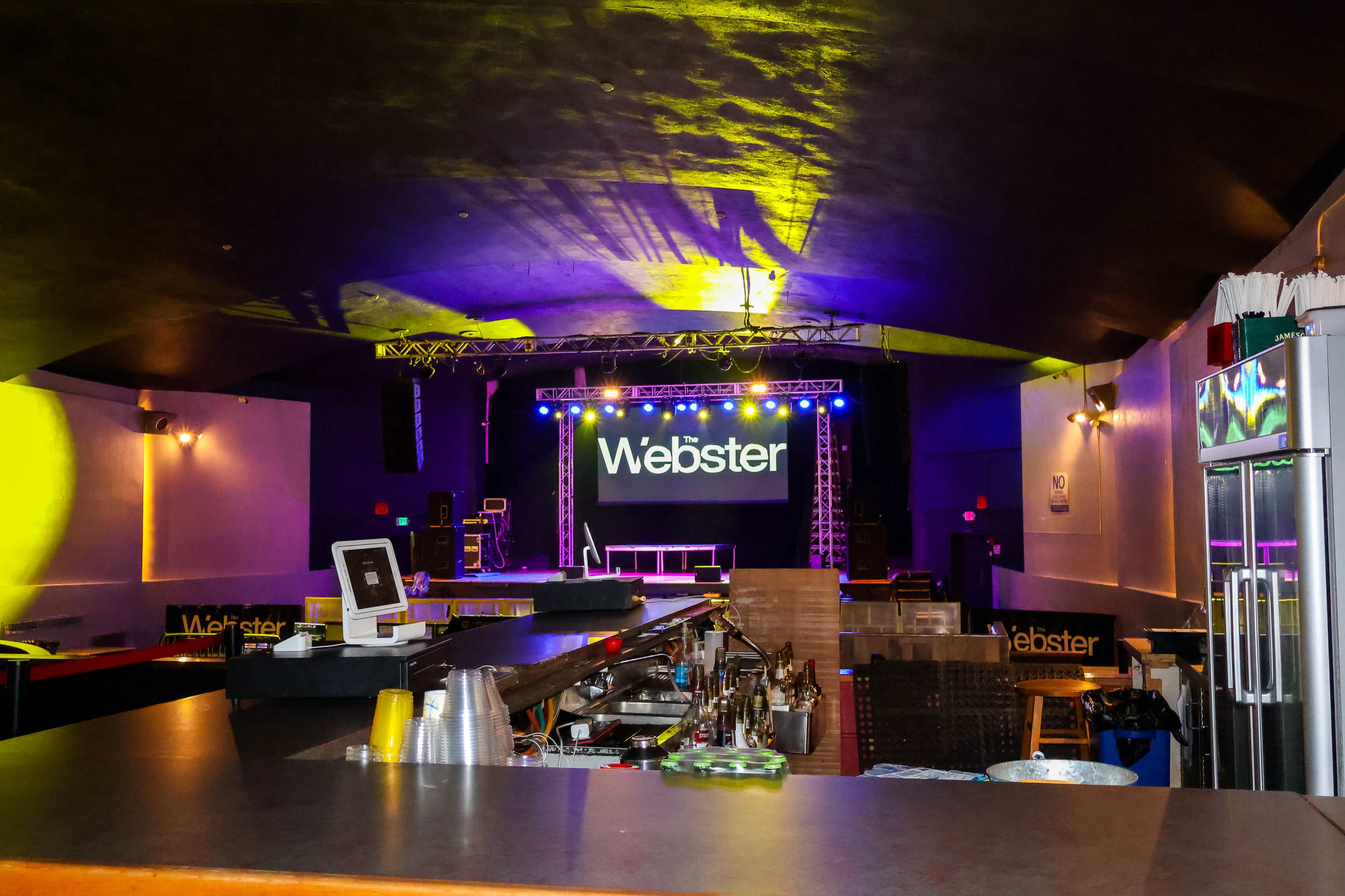The image shows an empty nightclub interior with a bar area in the foreground and a brightly lit stage featuring the name "Webster" in the background.