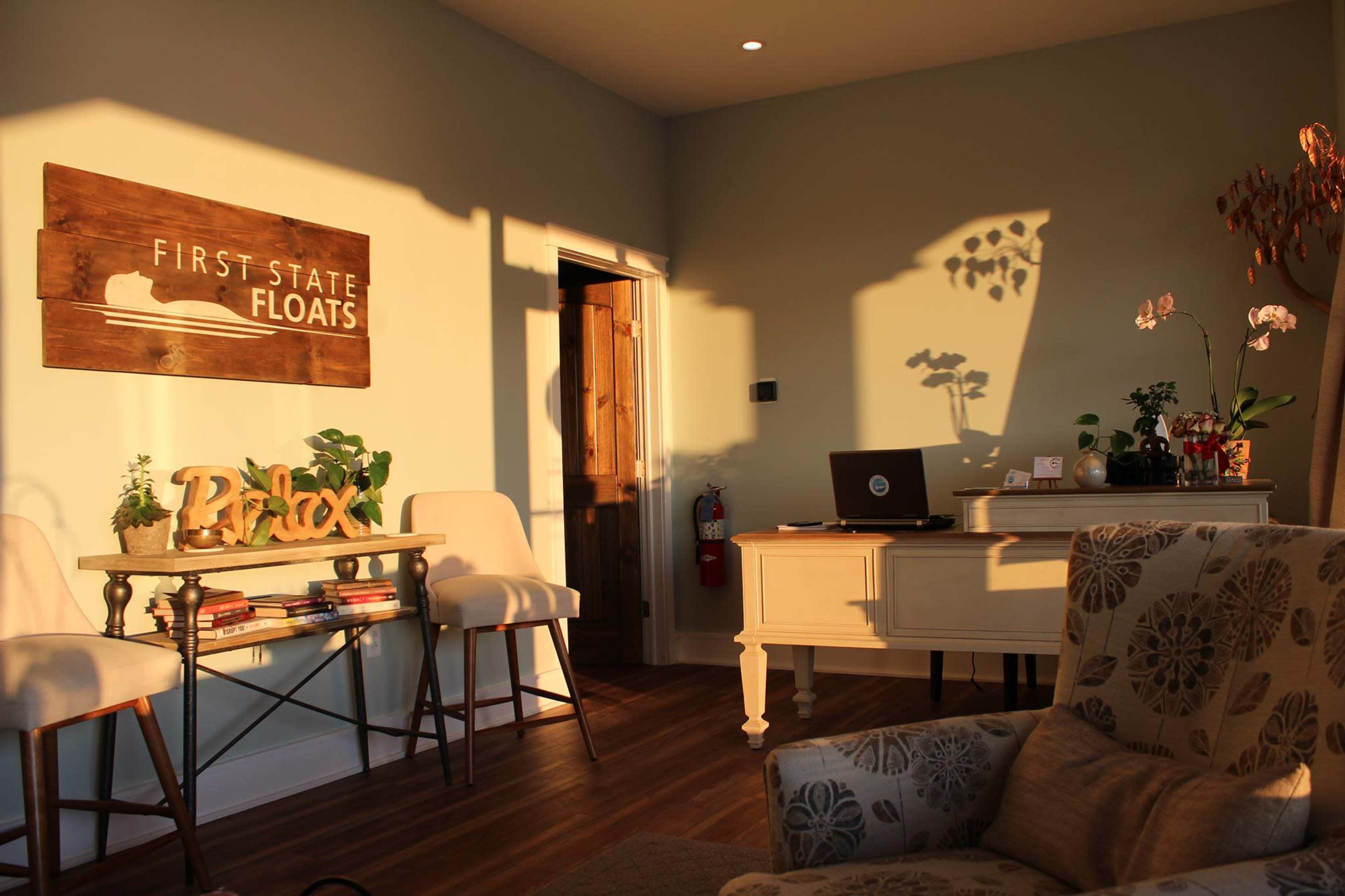 An inviting interior space with a wooden sign reading "FIRST STATE FLOATS," a desk with a laptop, and houseplants positioned around a cozy seating area.