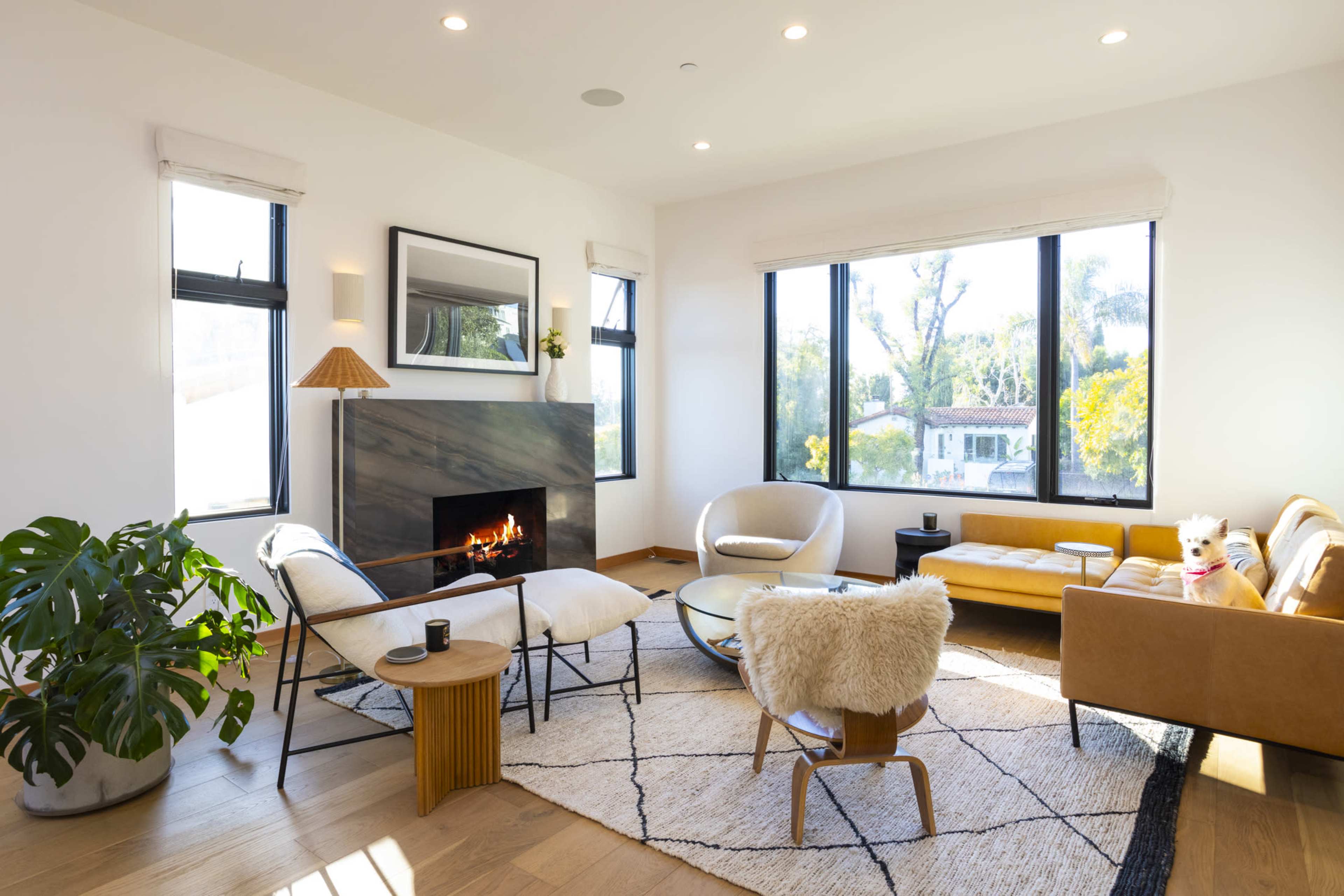 A modern living room features a fireplace, large windows, a couch, and two chairs arranged around a coffee table on a patterned rug.
