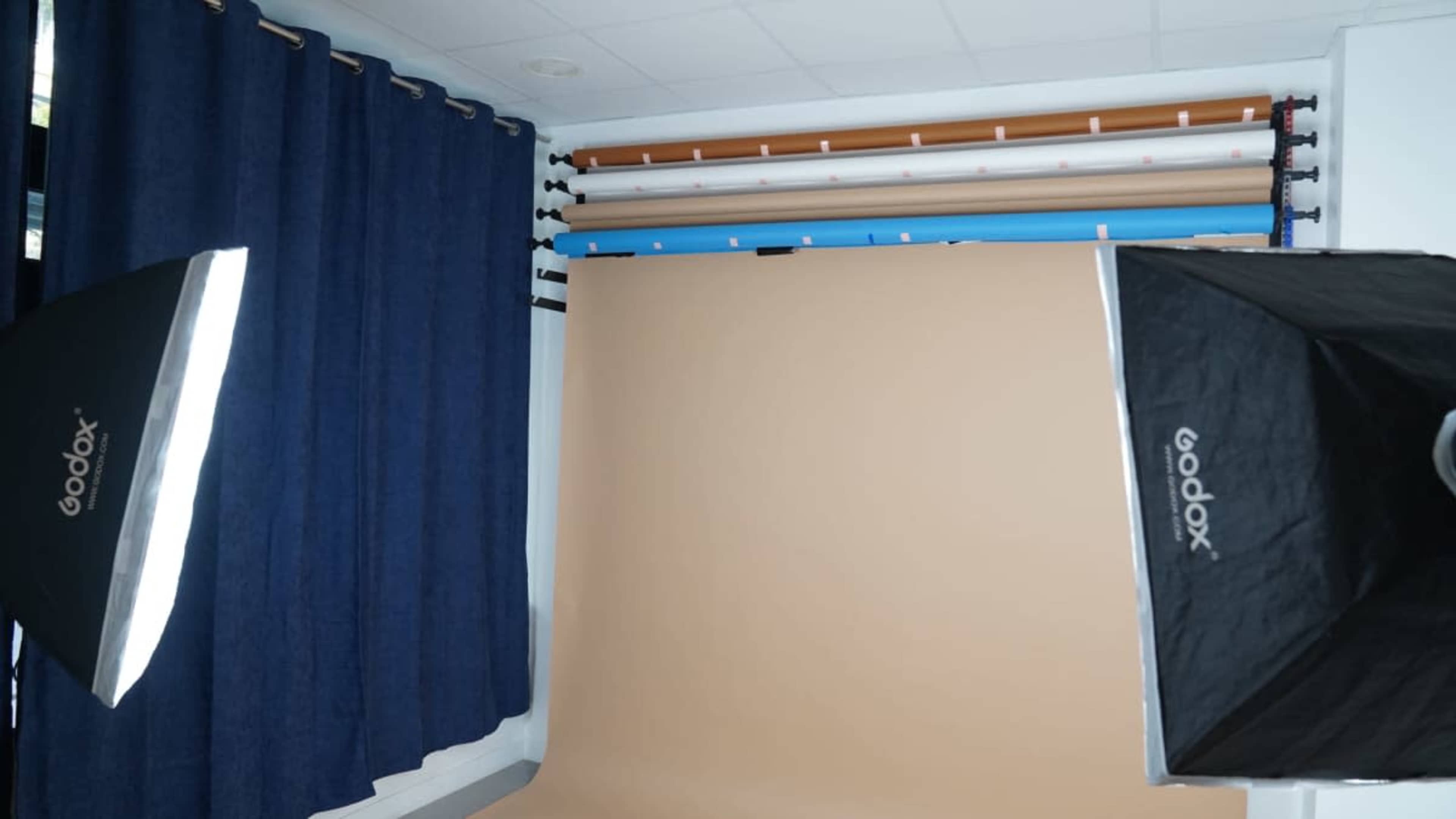 The image shows a photography studio setup with a brown backdrop, softbox lights, and curtains, alongside rolled backgrounds in various colors.