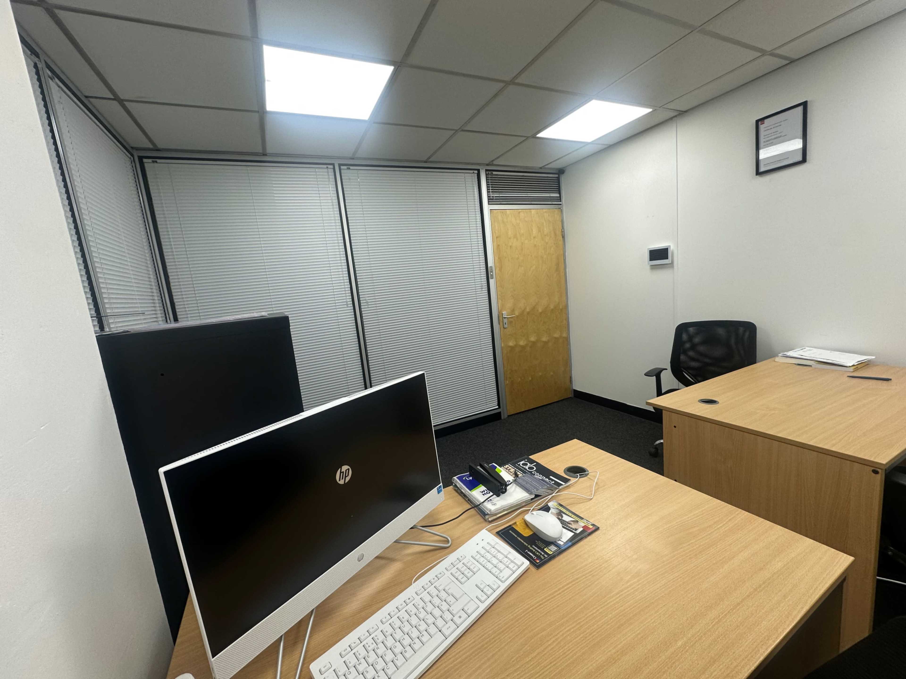 The image shows an office space with a wooden desk, an all-in-one computer, a black office chair, and sliding blinds on one side.