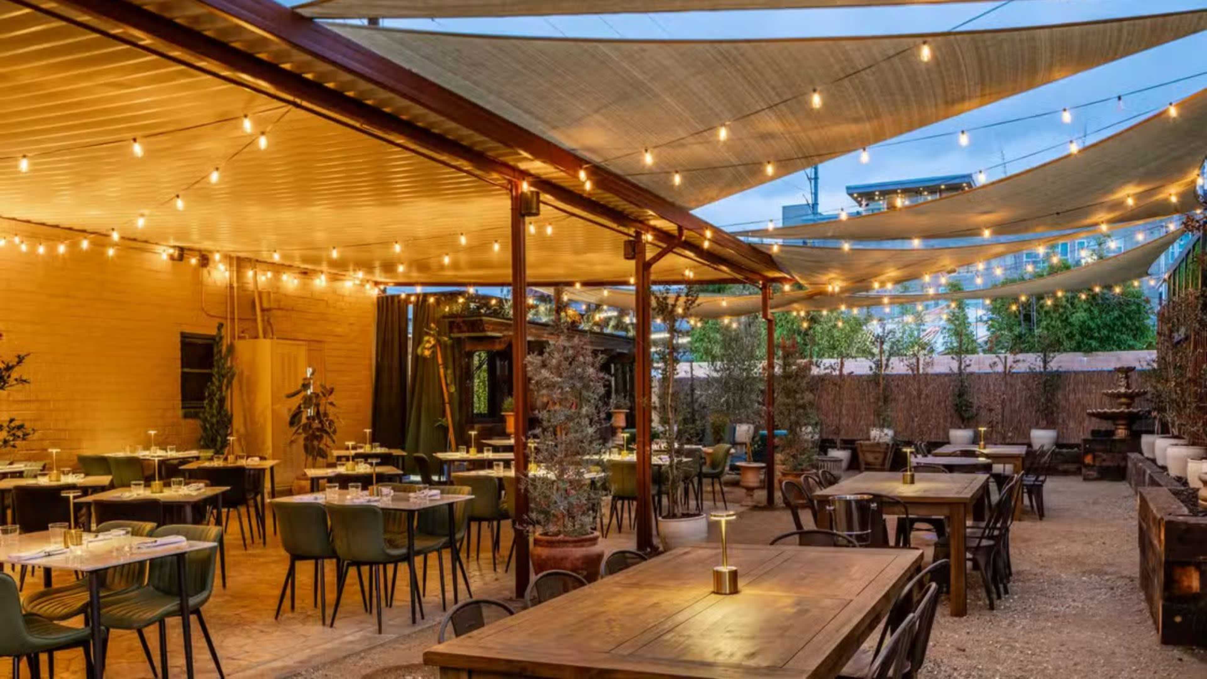 The outdoor dining area features wooden tables, green chairs, and string lights under a shaded canopy.