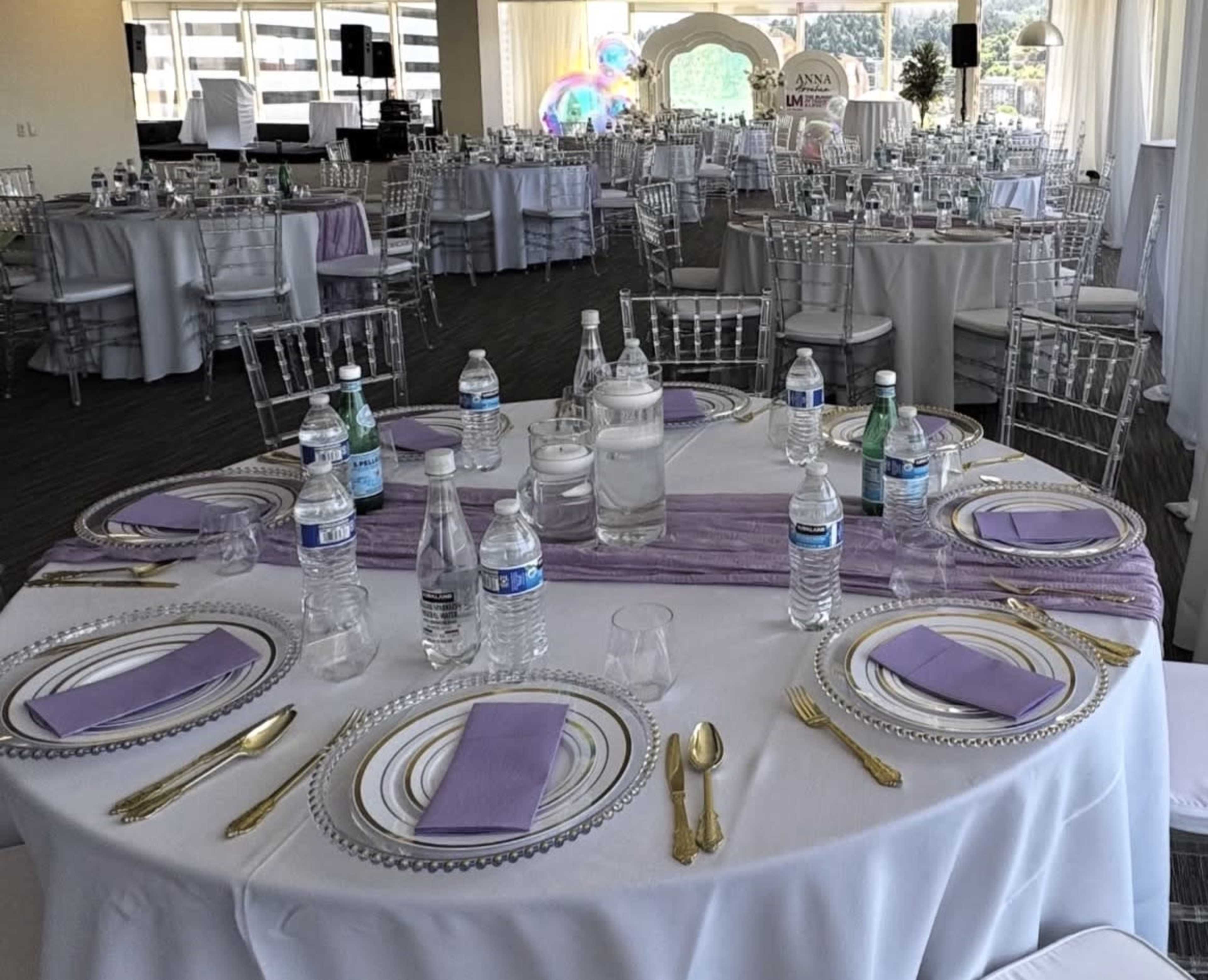The image shows a set dining table with clear plates, purple napkins, and multiple bottled waters, arranged in a banquet hall filled with covered tables and a white backdrop.