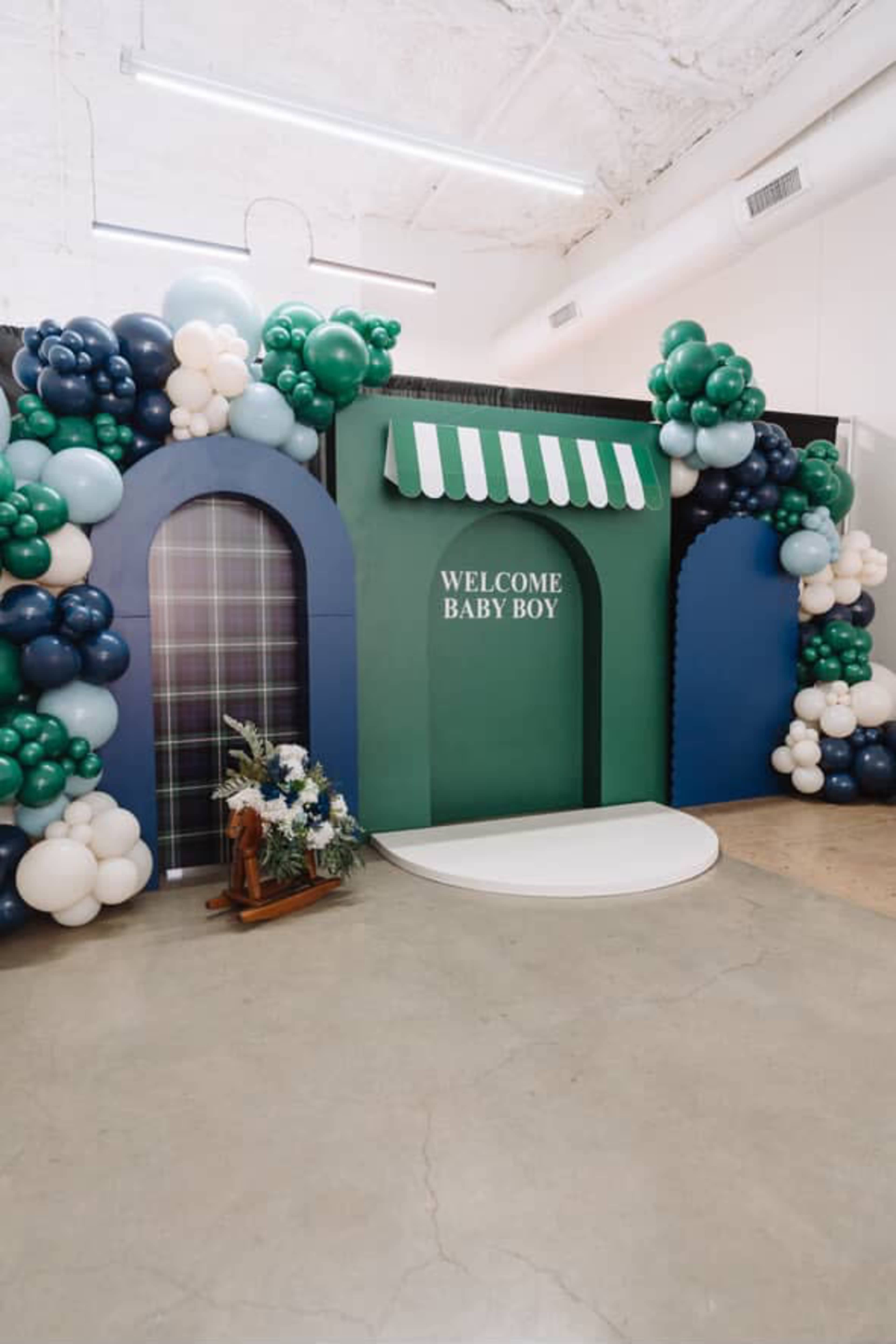 The image shows a decorated backdrop for a baby shower with a "Welcome Baby Boy" sign, featuring arches, balloons, and a wooden rocking horse.