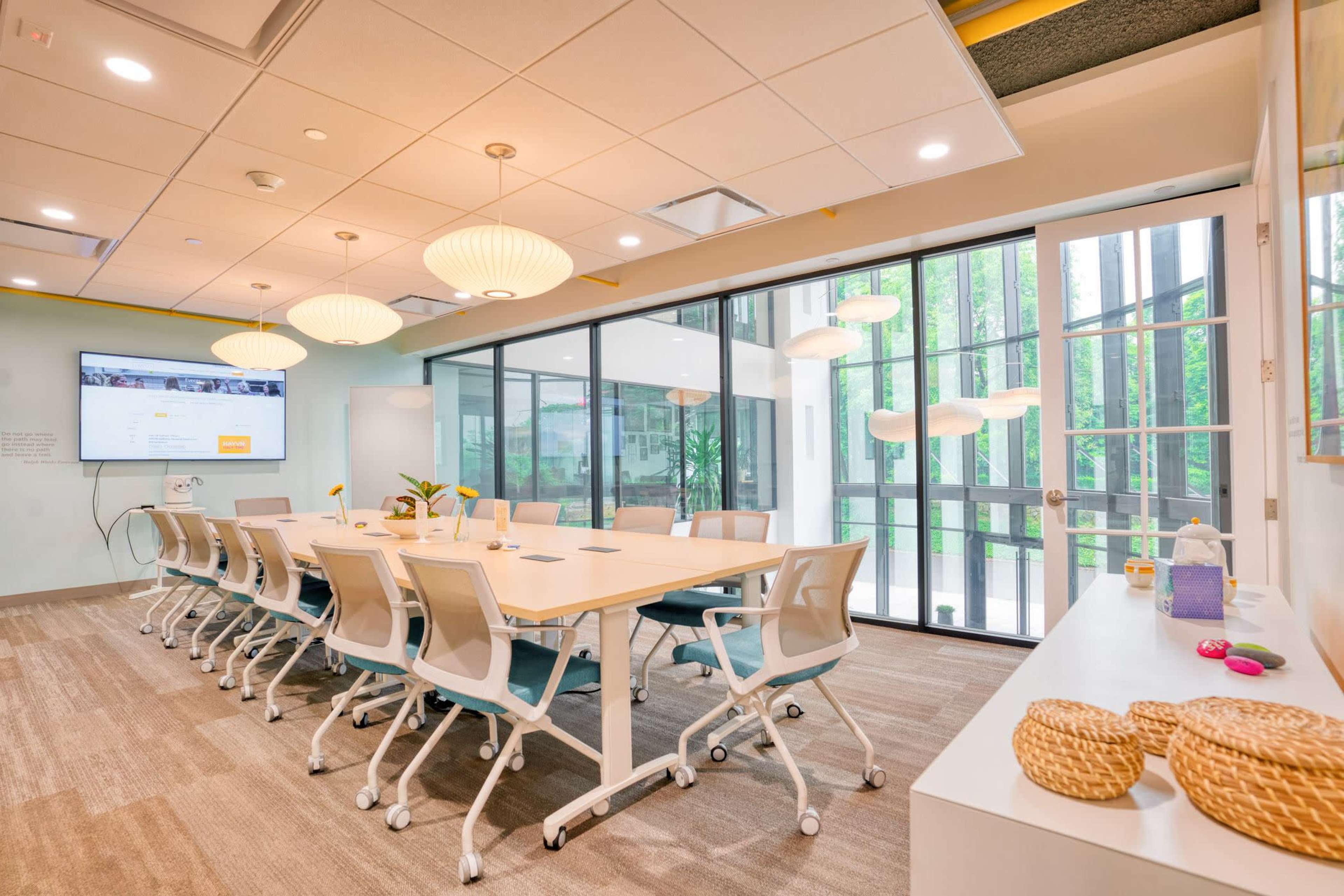 A modern conference room features a large white table surrounded by ergonomic chairs, with floor-to-ceiling windows overlooking greenery and a presentation screen on the wall.