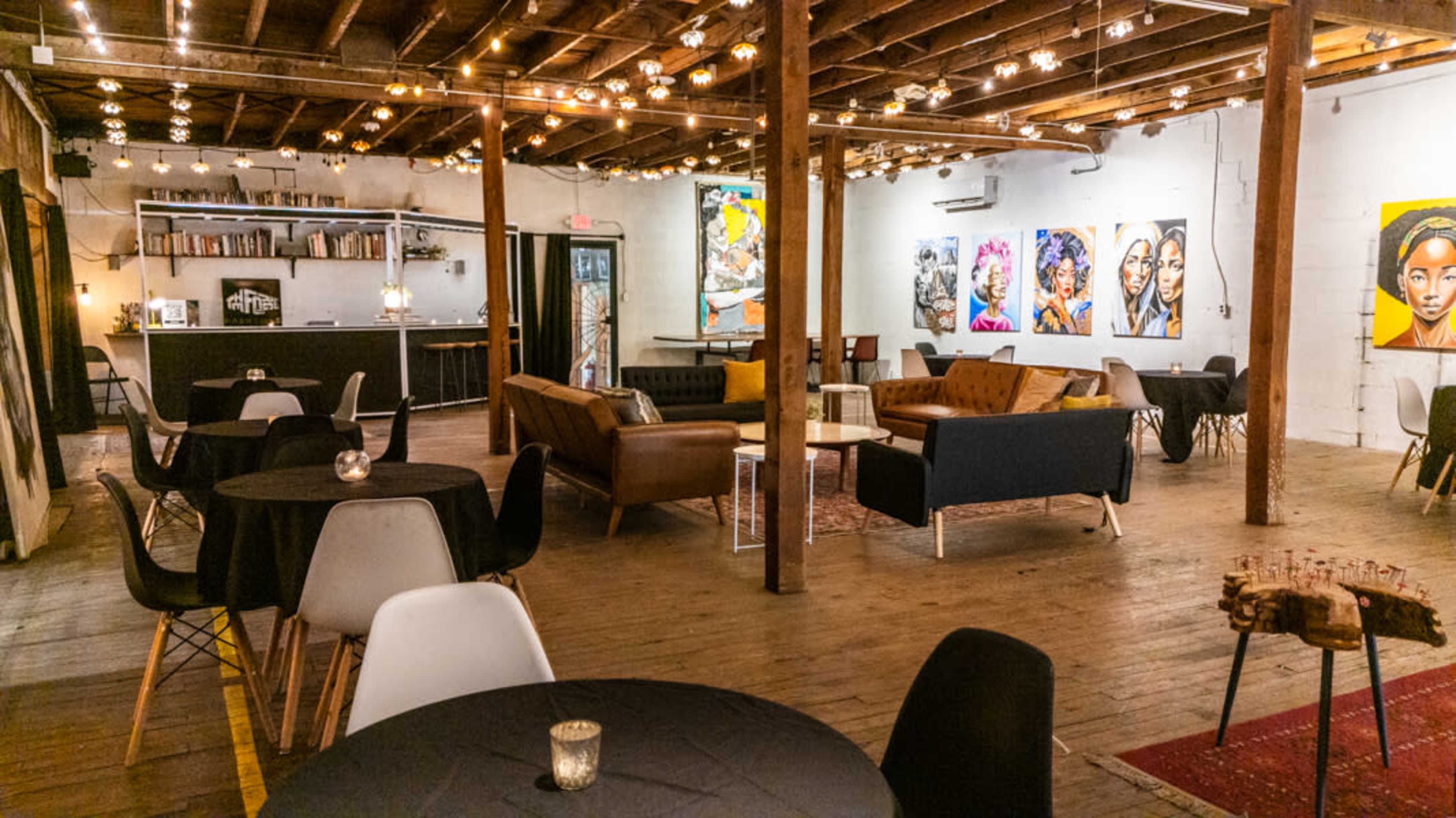 The image depicts a spacious indoor venue featuring wooden floors, exposed beams, and various seating arrangements, with artwork displayed on the walls.