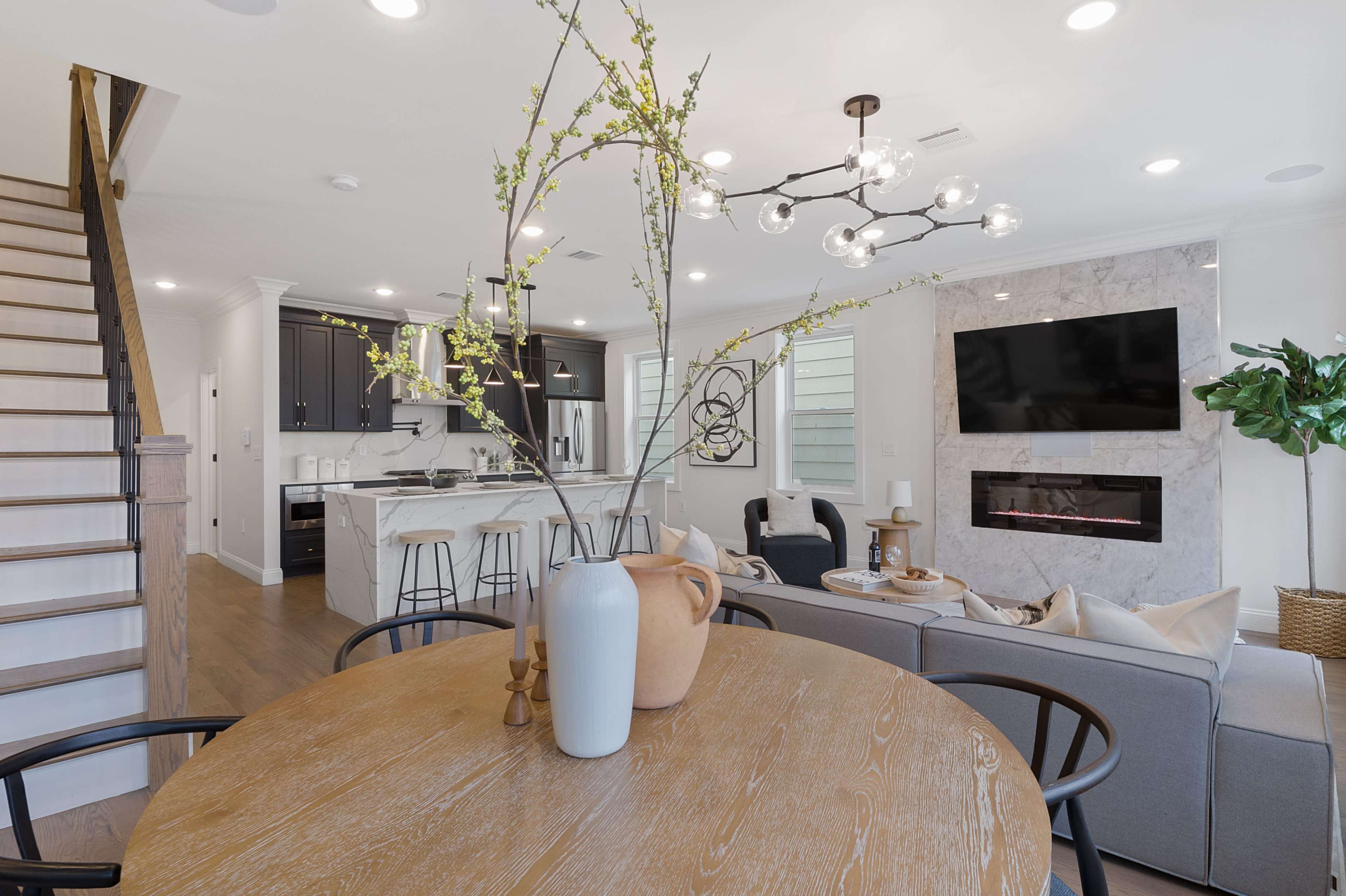 A modern living space with a dining area featuring a round wooden table, a cozy seating area with a fireplace, and a staircase leading to the upper level.