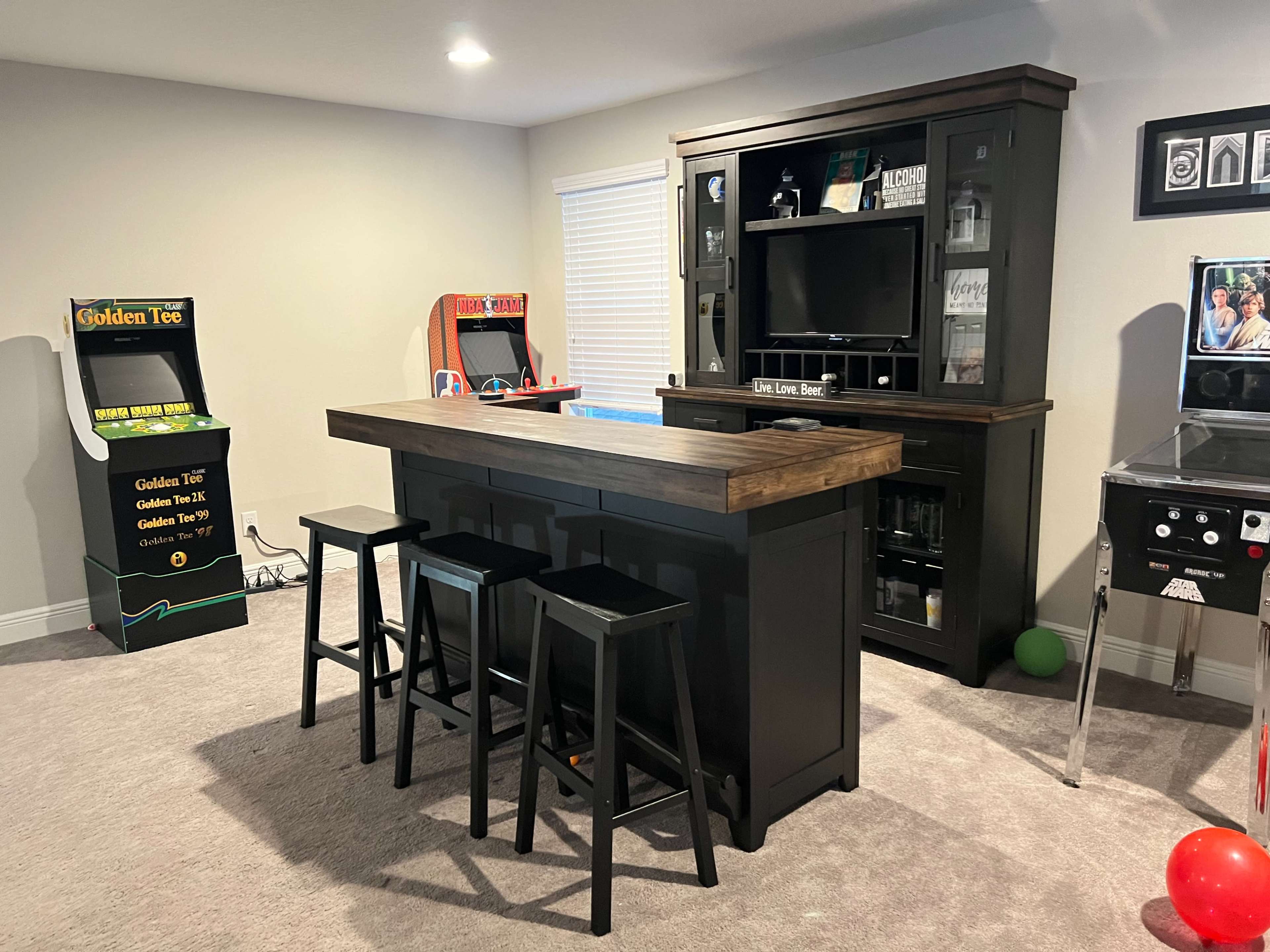 A home entertainment area featuring a bar with stools, a black cabinet holding a television, and two arcade machines in the background.