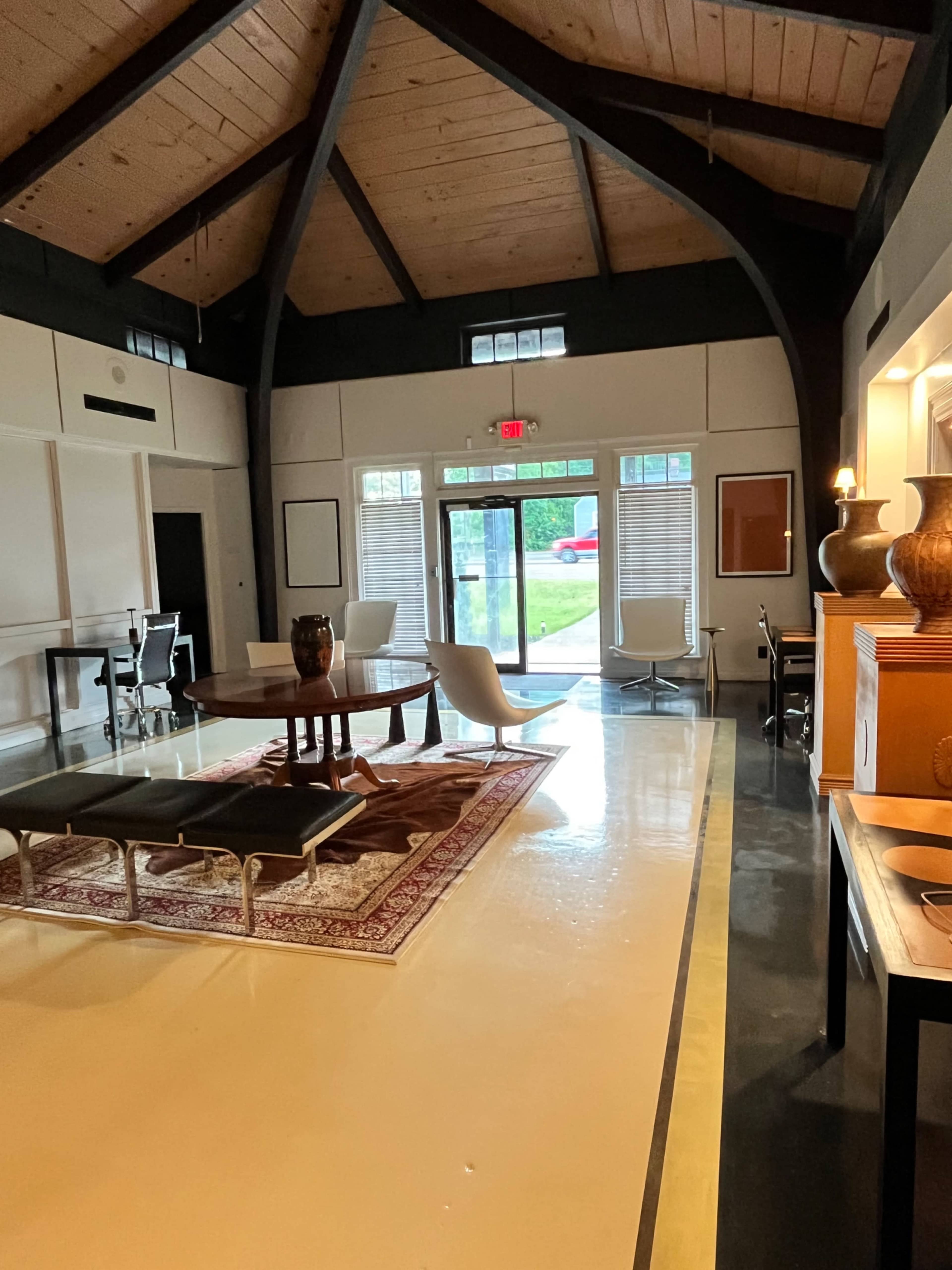 The interior of a modern communal lounge features a round table, a large area rug, and various seating arrangements under a high, exposed beam ceiling.