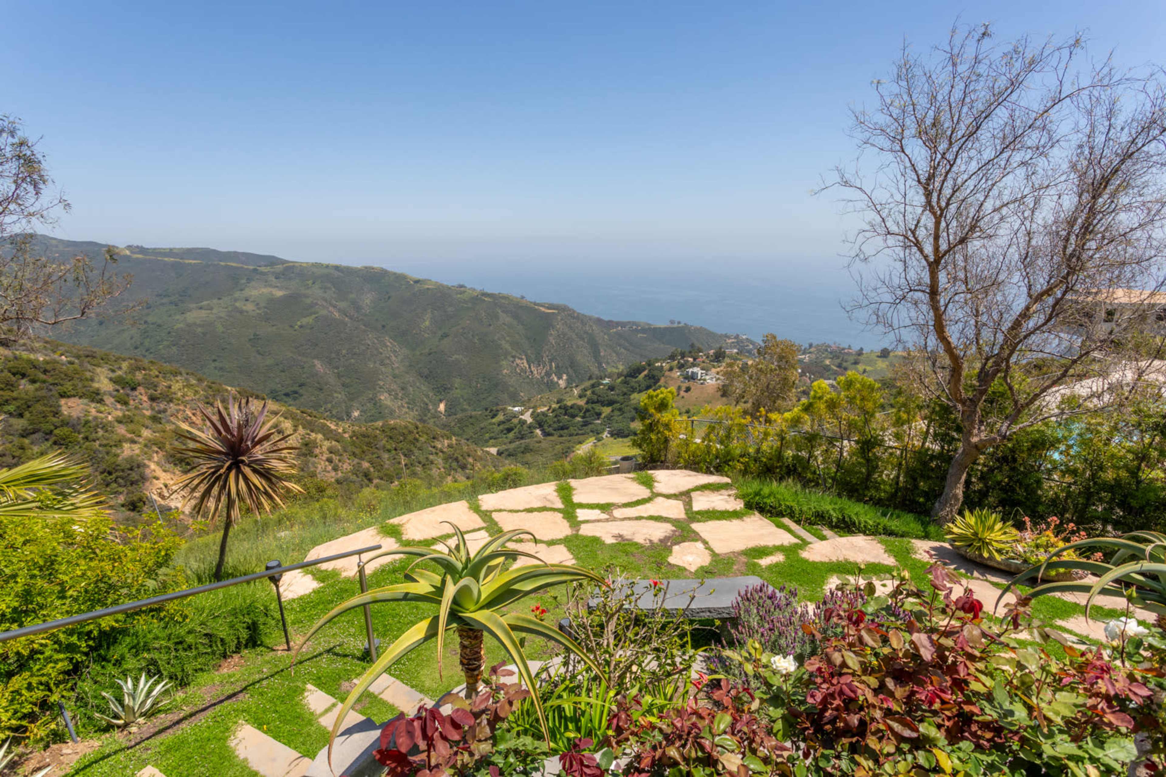 A landscaped garden with various plants and stone pathways overlooking a green hillside and the ocean in the distance.