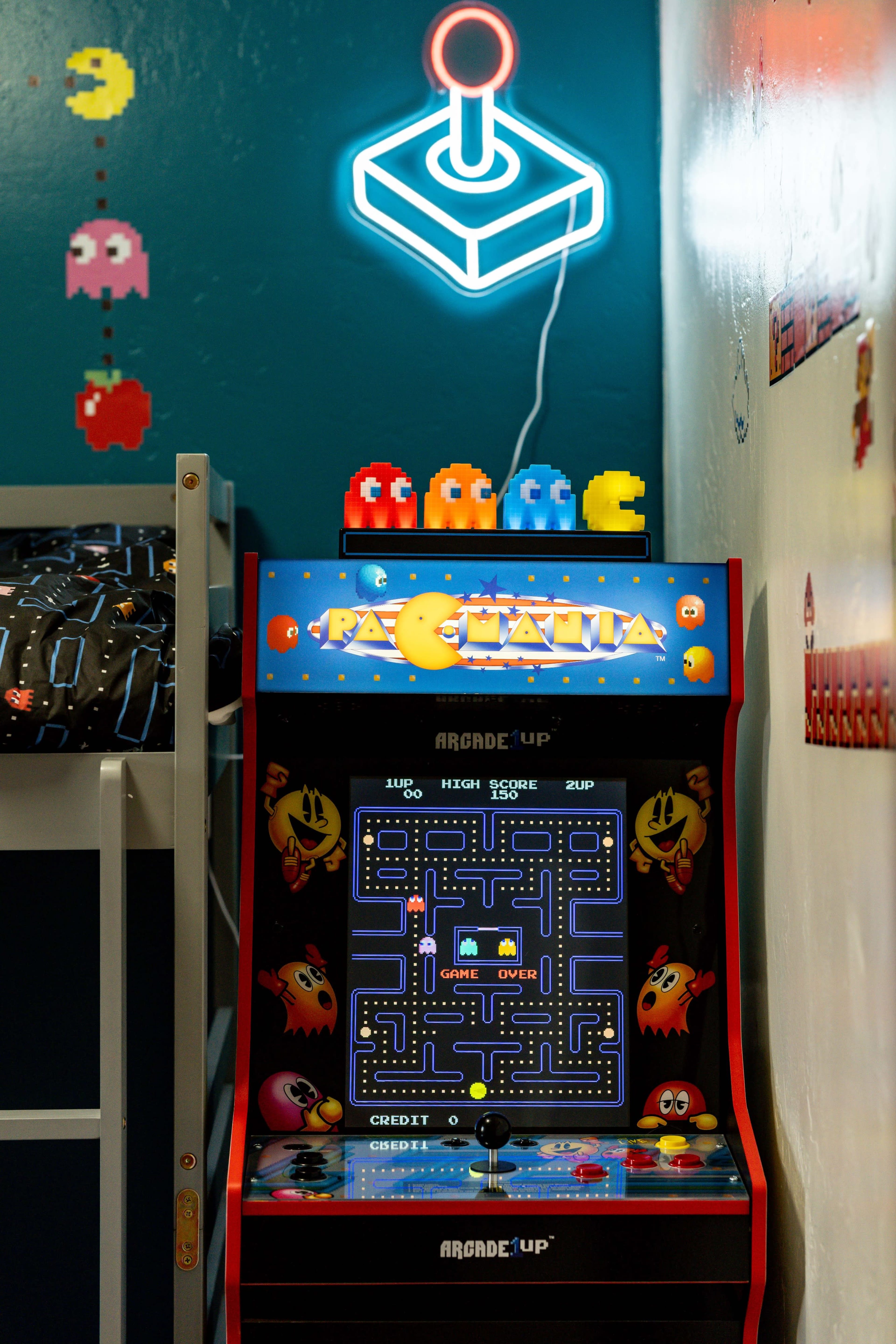 A vintage Pac-Man arcade game machine located against a blue wall with a neon joystick light above it and colorful ghost decorations nearby.
