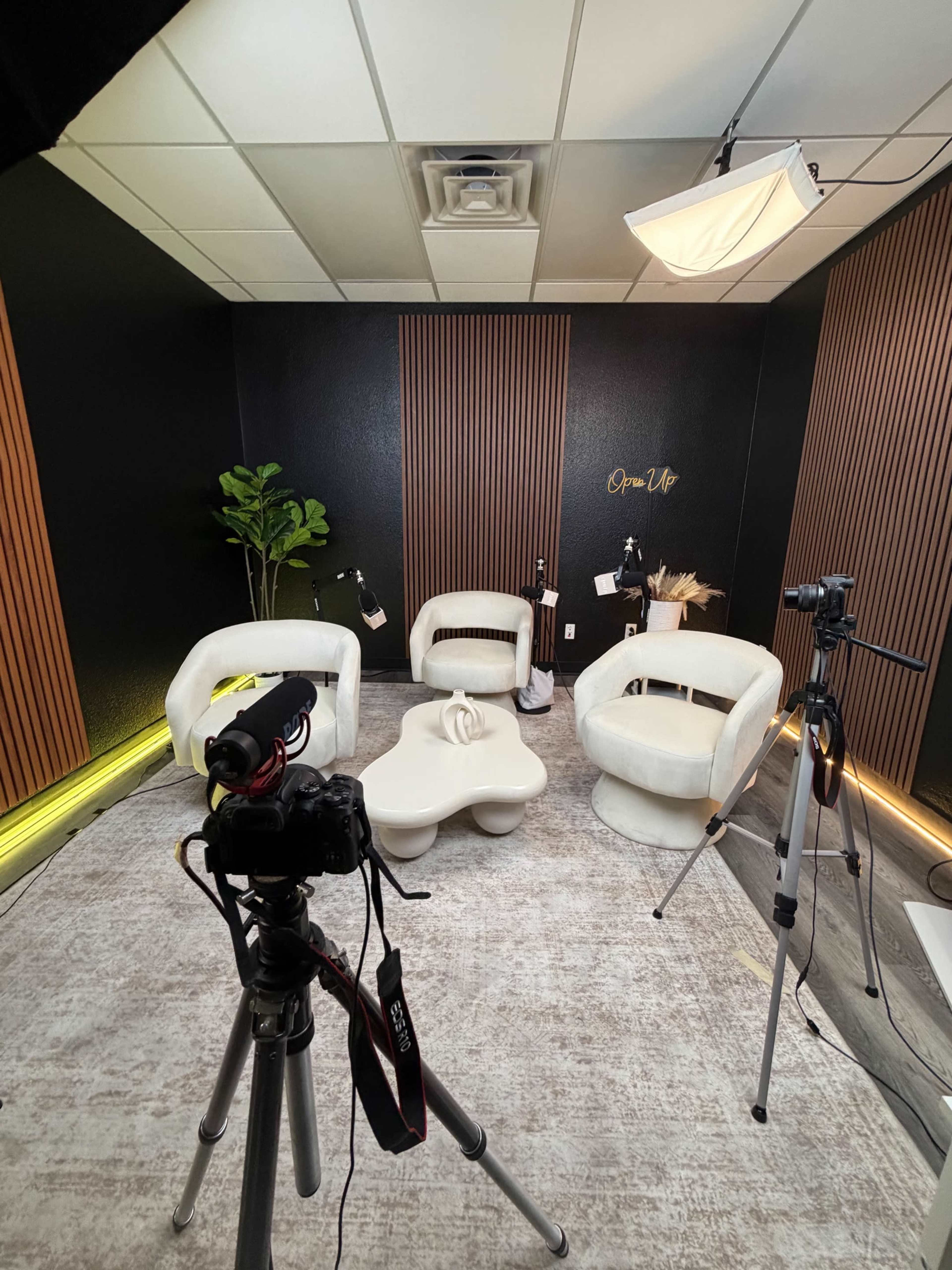 The image shows a modern filming studio setup with two white chairs, a white coffee table, plants, and camera tripods positioned around the room.