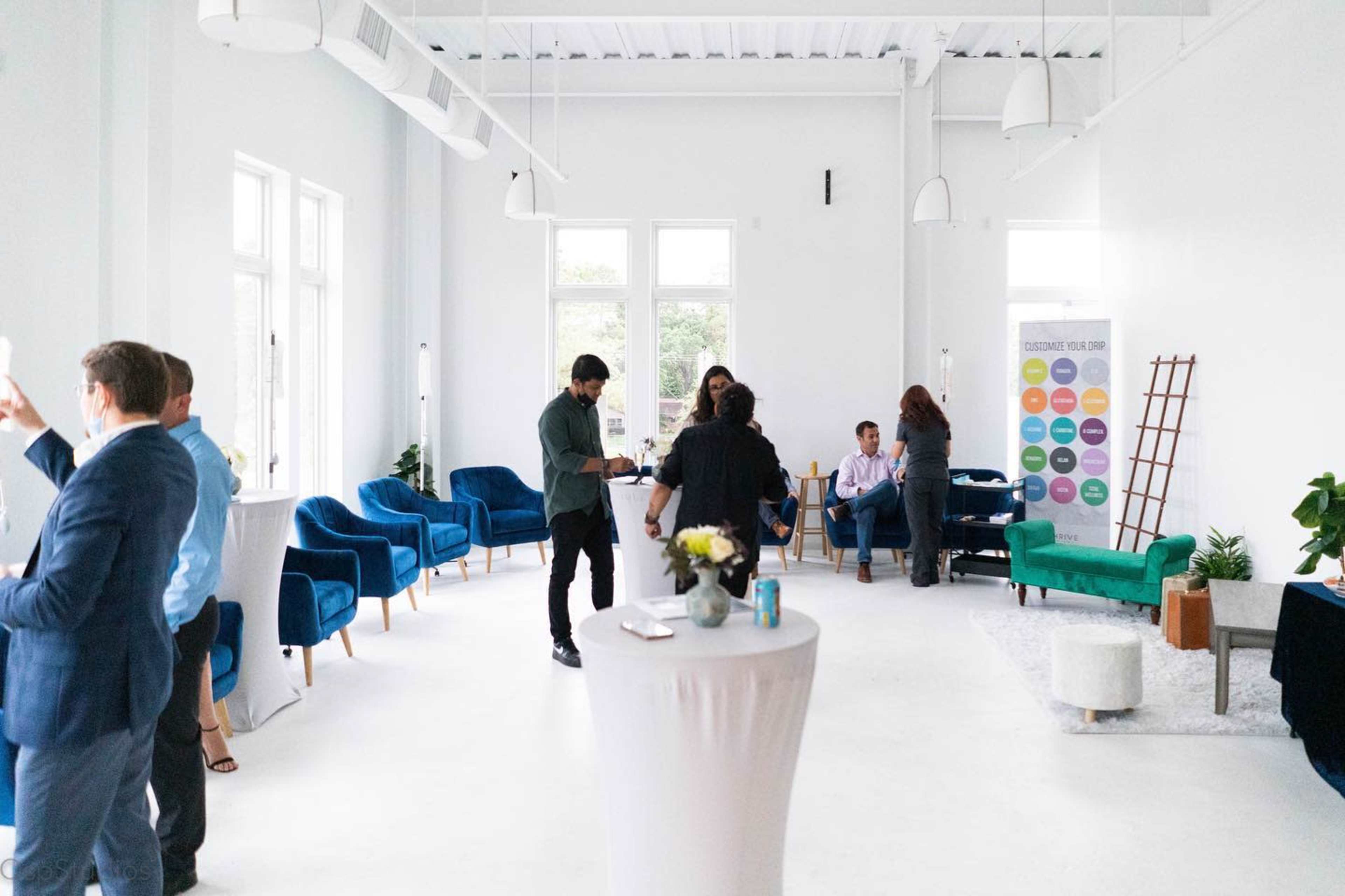 A bright, modern event space features people mingling around tables and plush blue chairs, with large windows allowing natural light to fill the room.