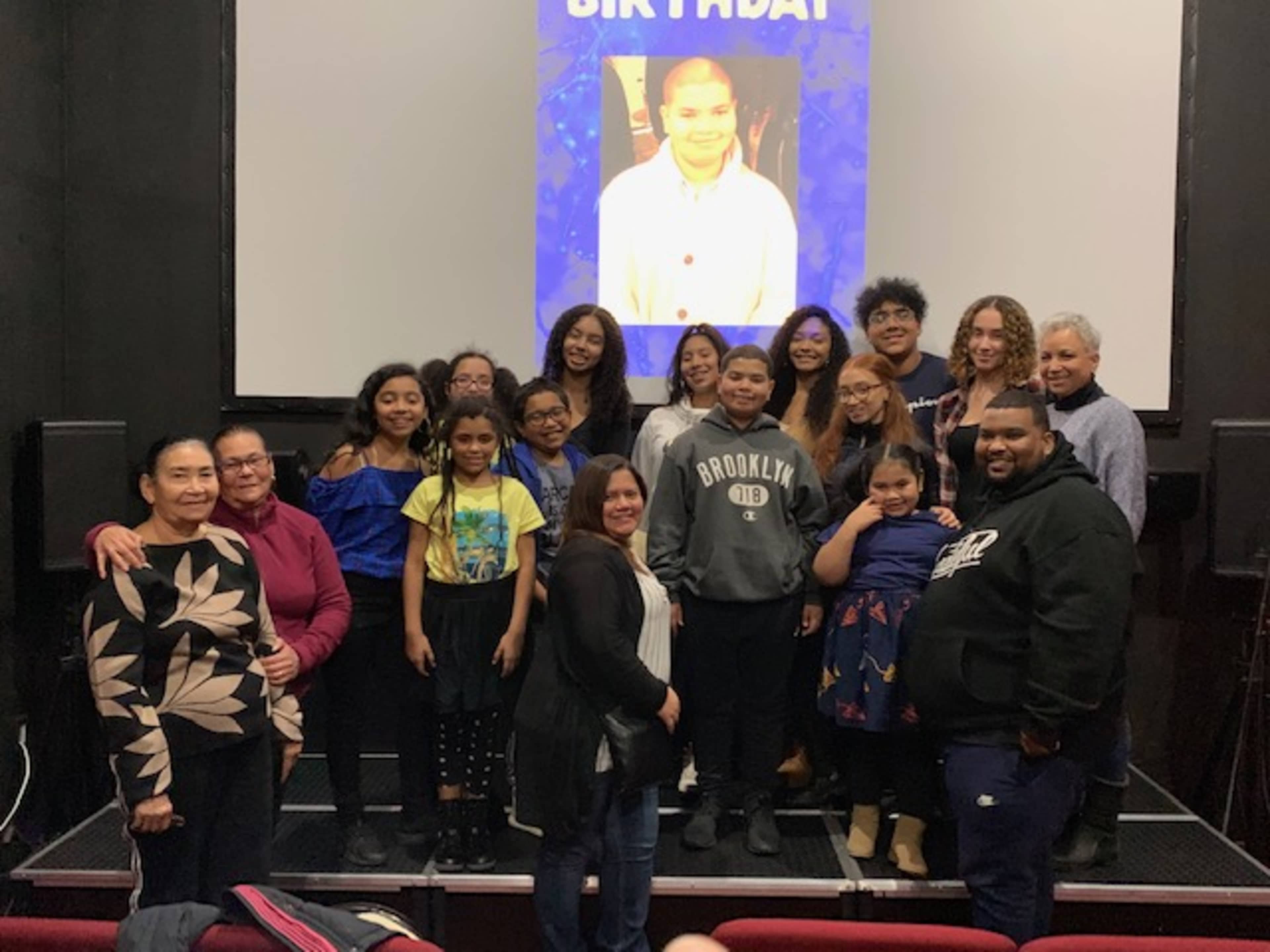 A group of people, mostly children and teenagers, gather for a birthday celebration in front of a projection screen.