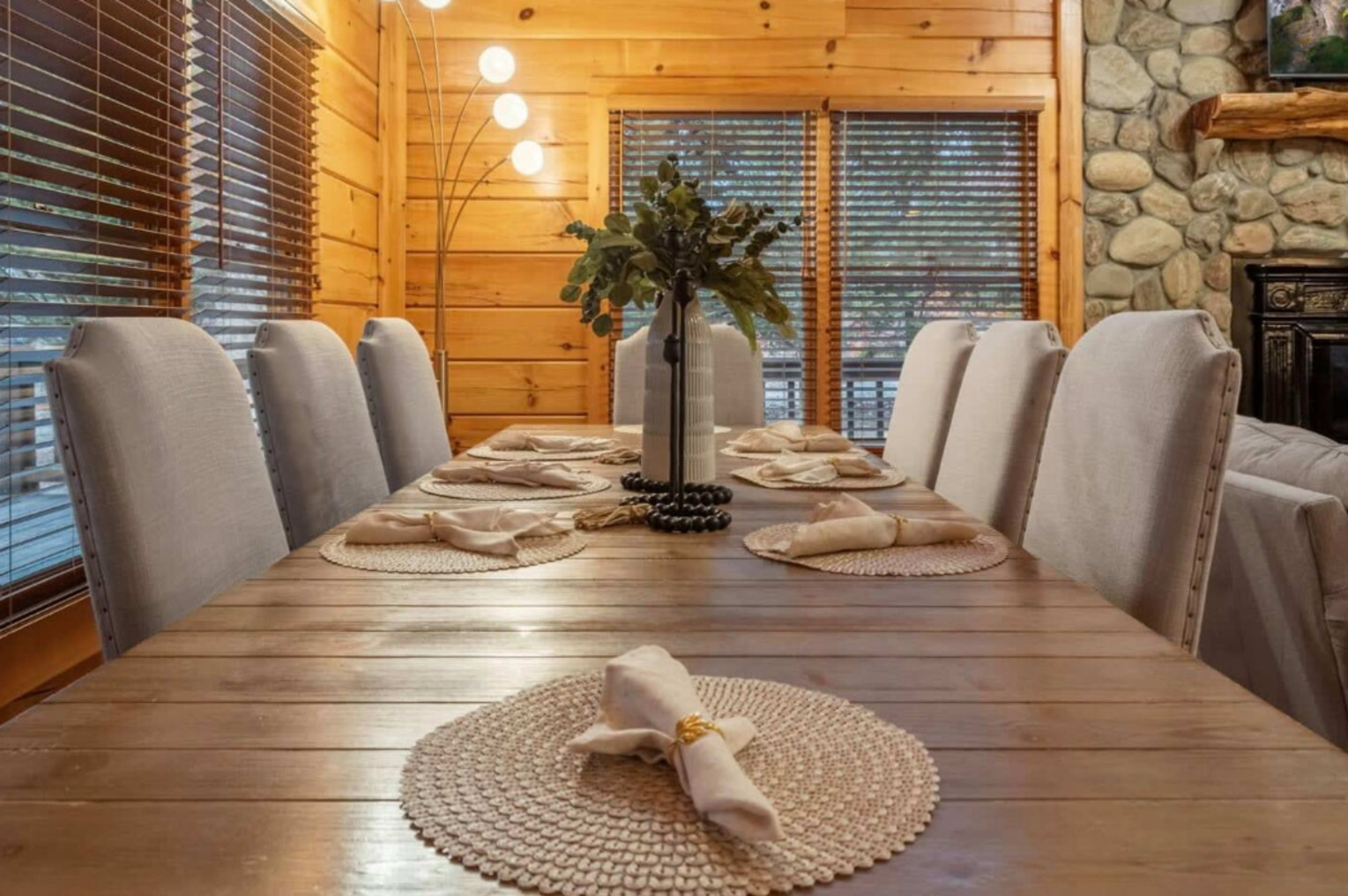 A long wooden dining table is set with placemats, napkins, and a vase of greenery in a room with wood paneling and large windows.